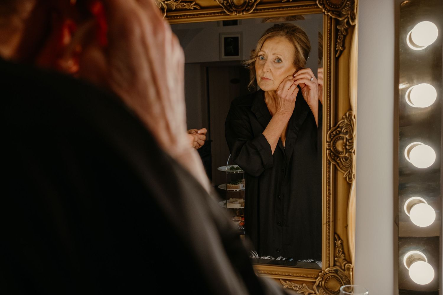 A reflection in an ornate mirror shows someone applying makeup near vanity lights.
