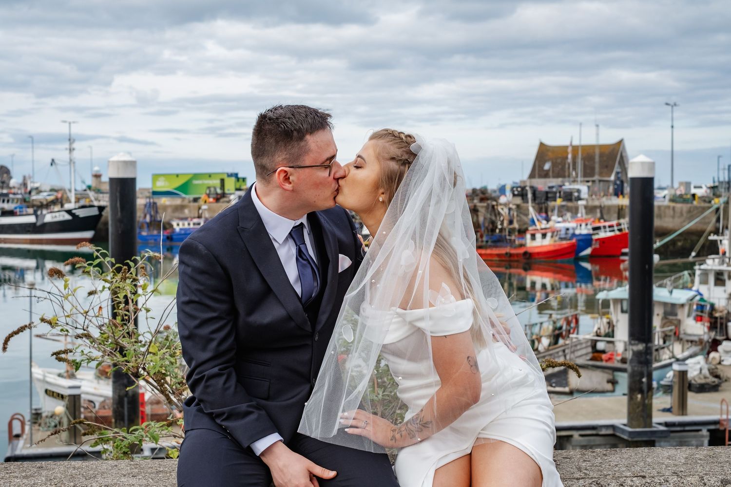 Bride and groom sharing a romantic kiss at a harbor, with colorful boats and cloudy sky in the background.
