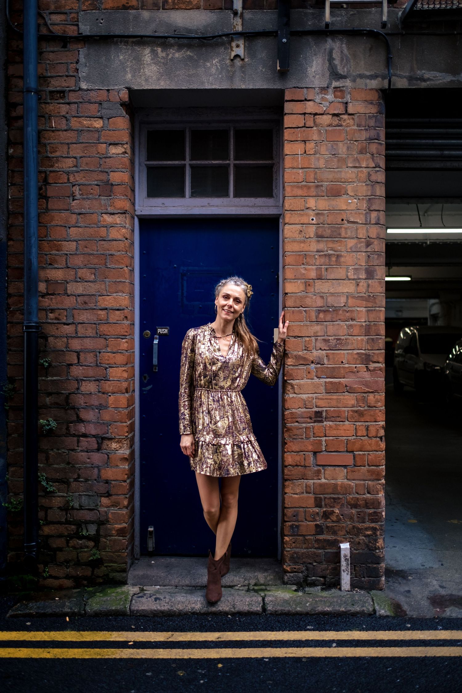 Woman in gold sequin dress posing by blue door on brick wall street.