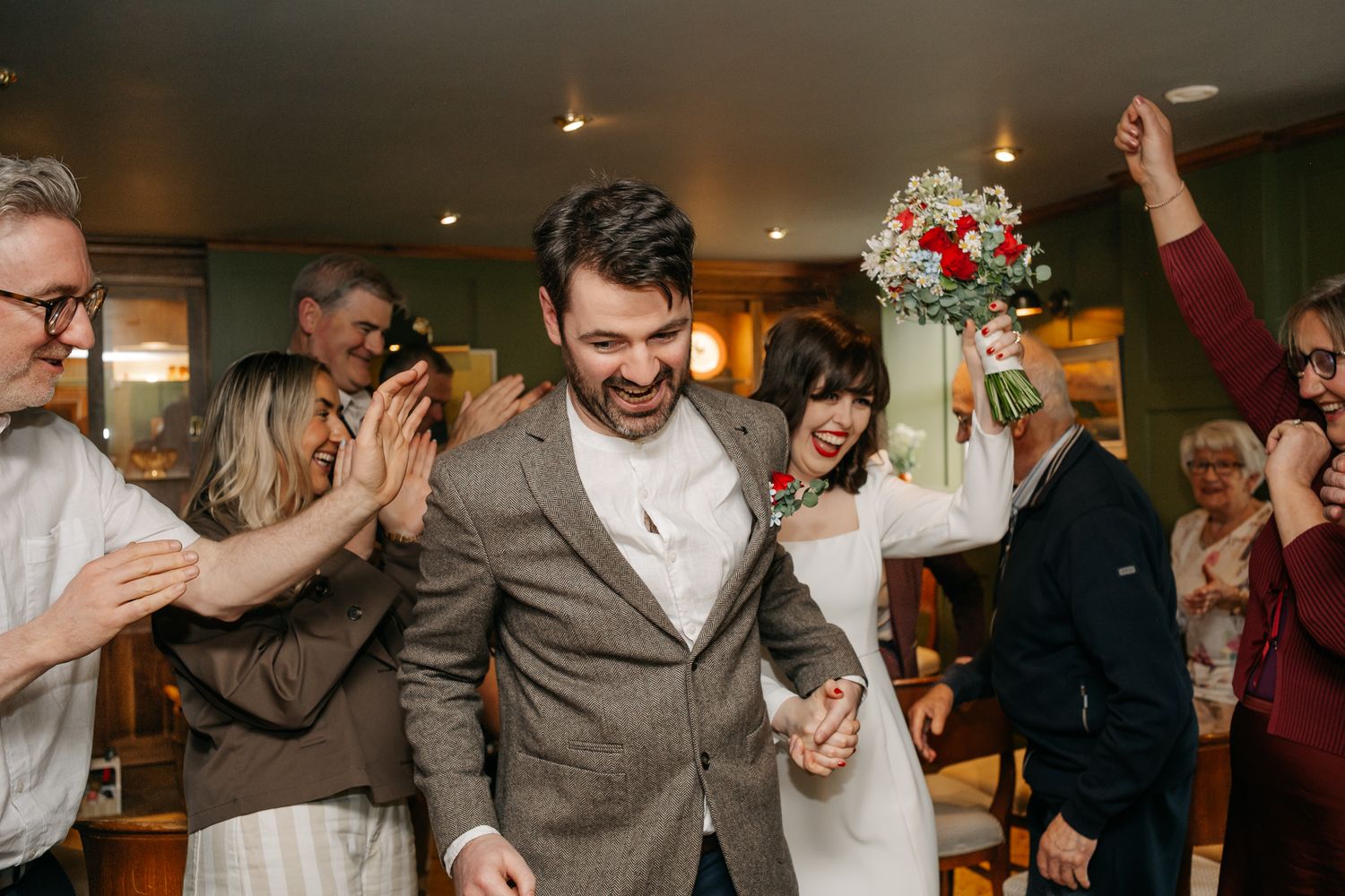 Joyful newlyweds walk through cheering guests, bride holding red and white bouquet aloft during wedding celebration indoors.