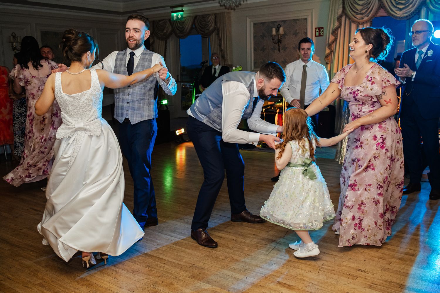 Wedding guests dancing joyfully on the dance floor, including a flower girl and groomsmen in grey vests.