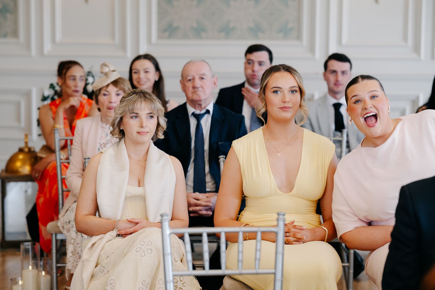 Wedding guests seated in elegant white chairs, smiling and reacting joyfully during an indoor ceremony.