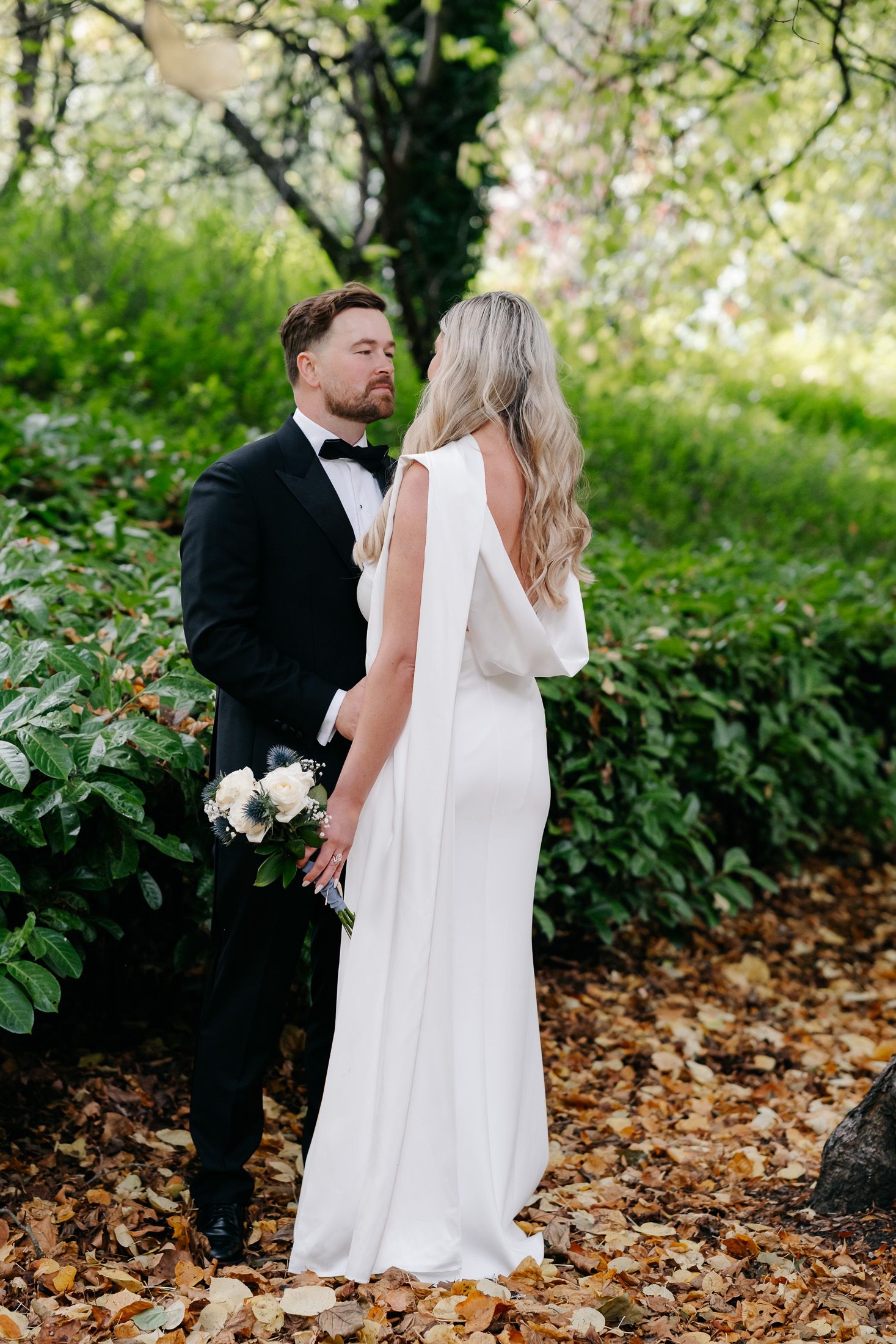 A bride in a white backless gown and groom in a black tuxedo share a romantic moment among autumn leaves.