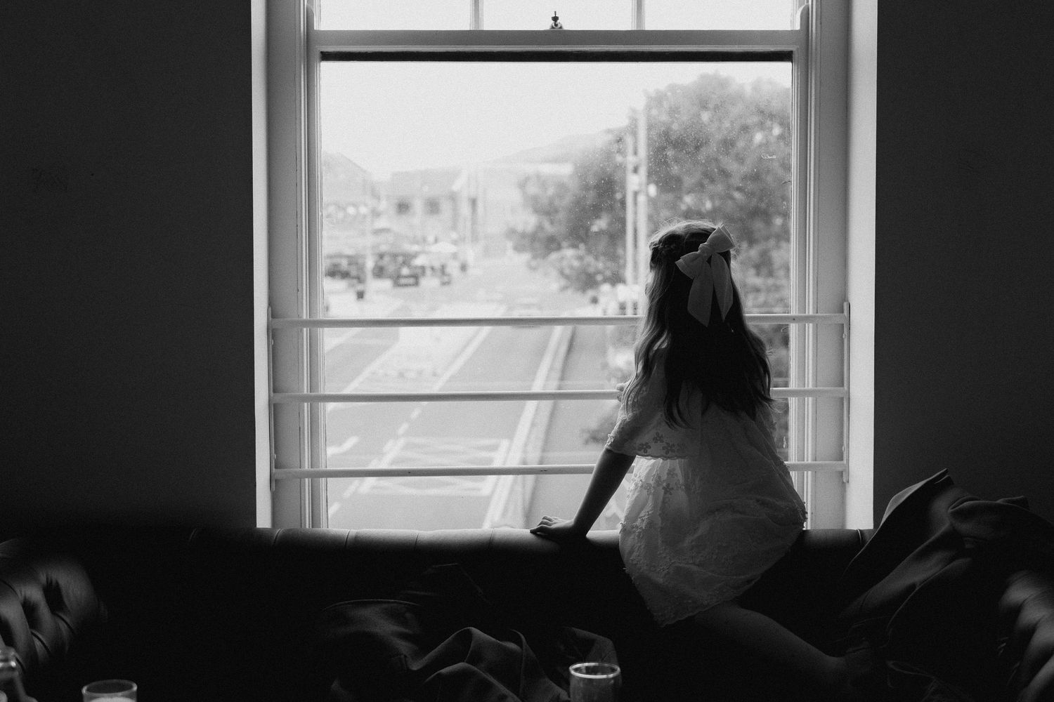 Black and white photo of a woman in a white dress sitting on a couch, gazing out a large window at a city street.