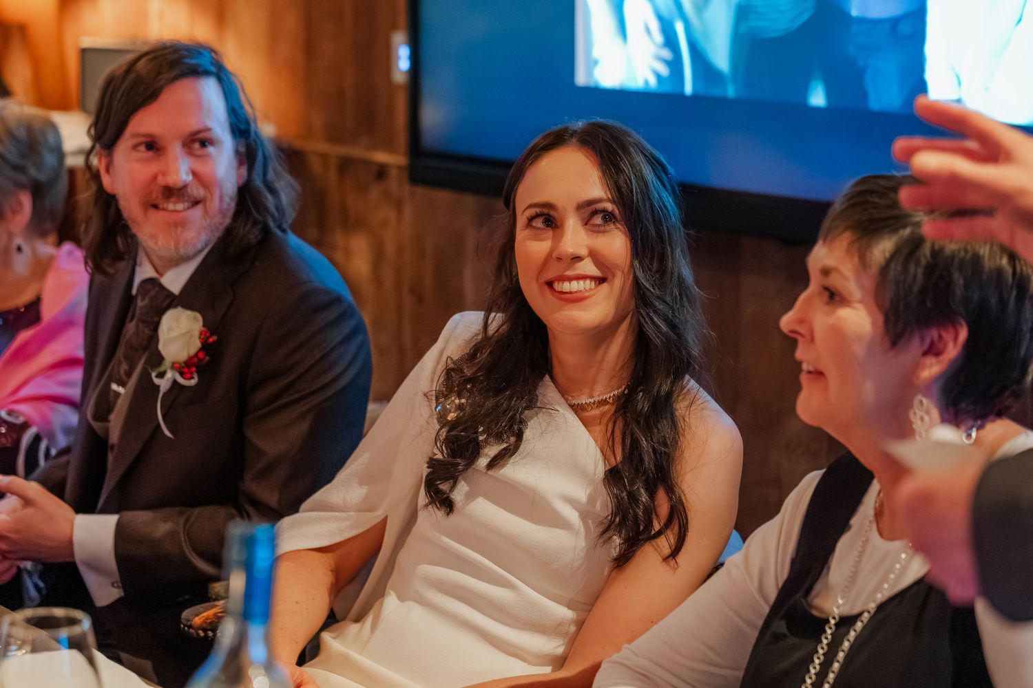 Bride in white dress laughing with groom and guest at wedding reception dinner table.