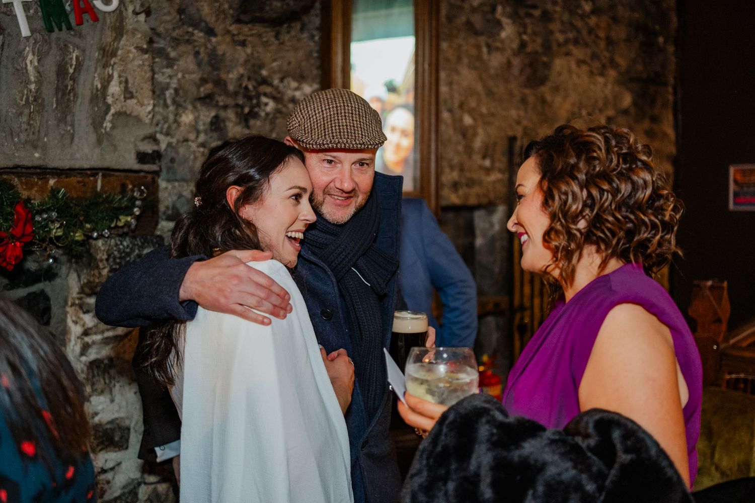 A man in a flat cap embraces a woman in white at a rustic stone-walled venue while another woman smiles nearby.