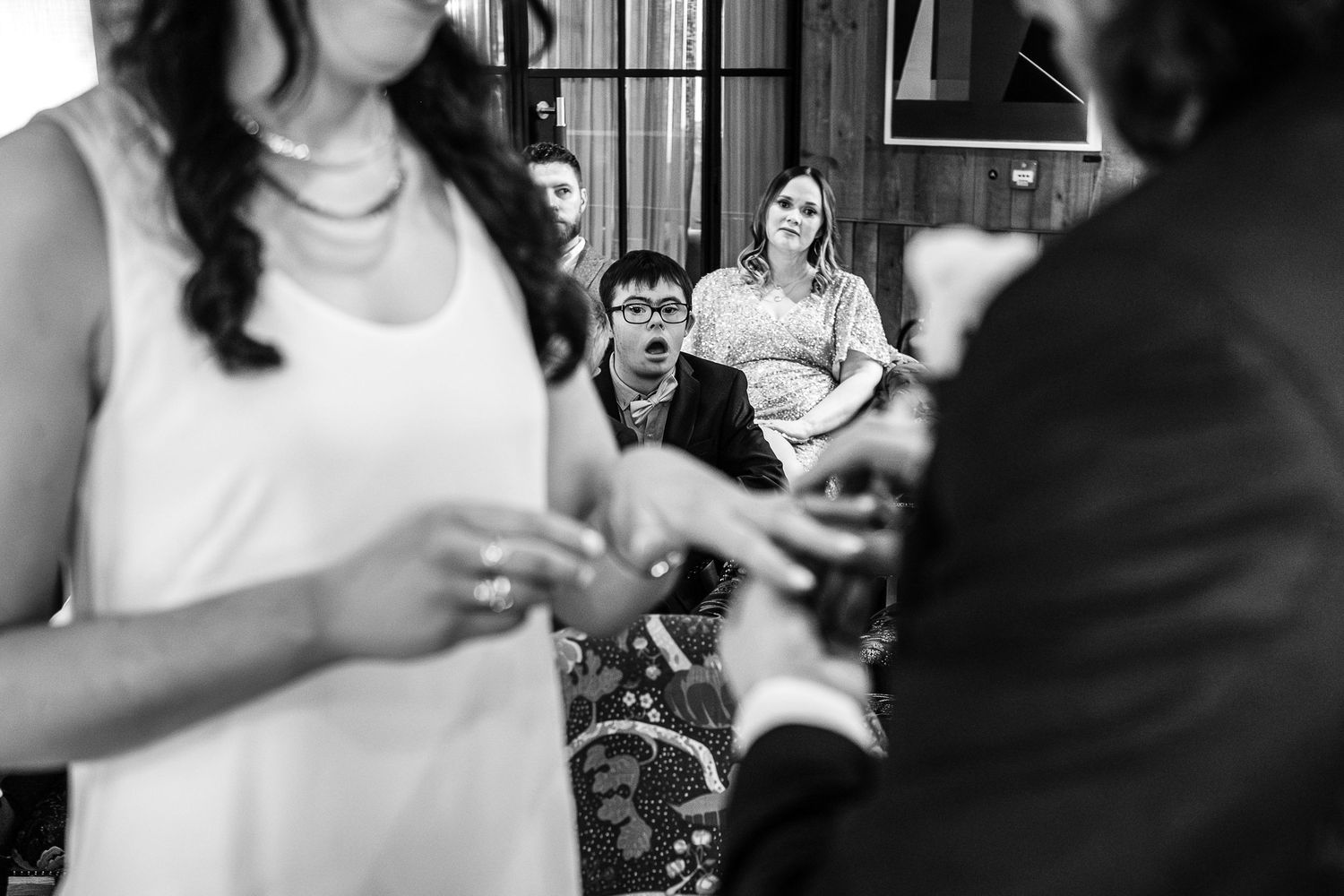 Black and white wedding ceremony photo showing guests watching emotional exchange of rings indoors.