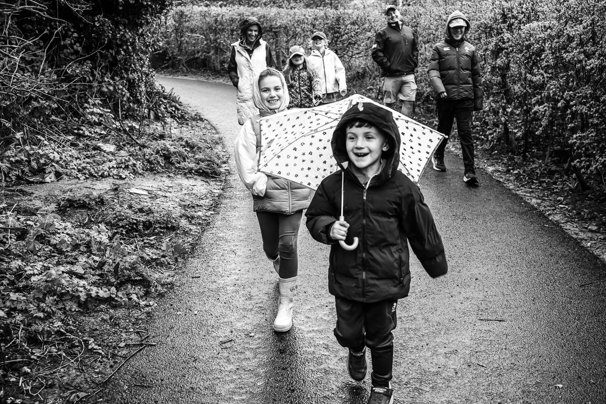 Black and white photo of joyful children running on a path with adults walking behind them outdoors.