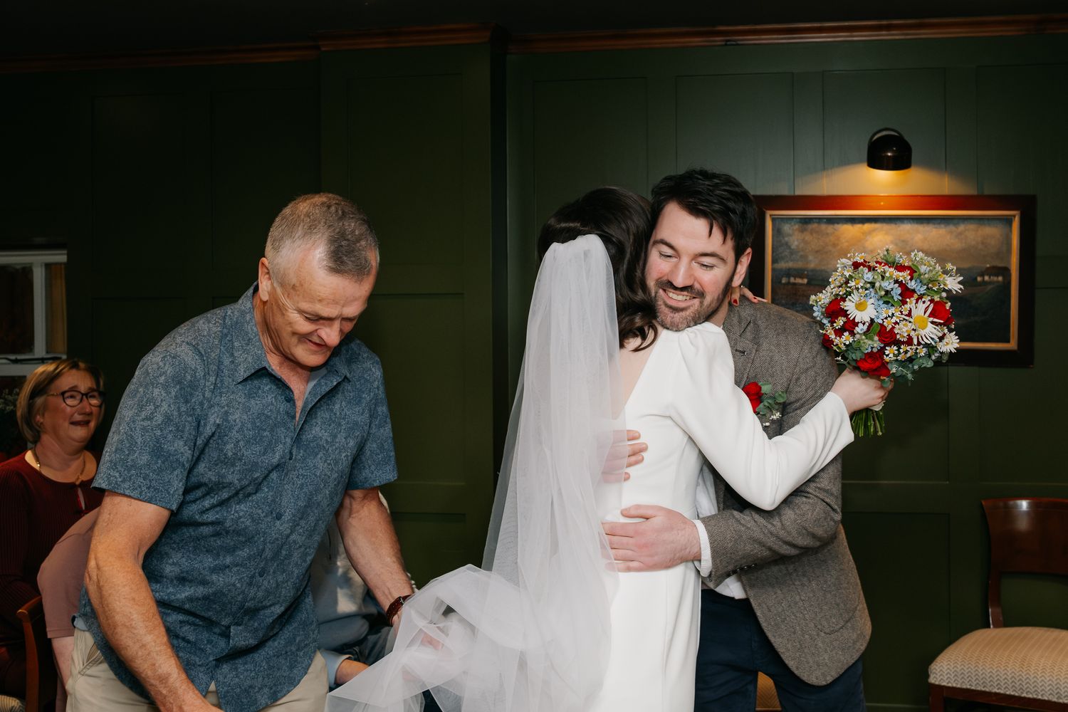 Bride in white dress and veil hugs groom in gray suit holding red bouquet at indoor wedding celebration.