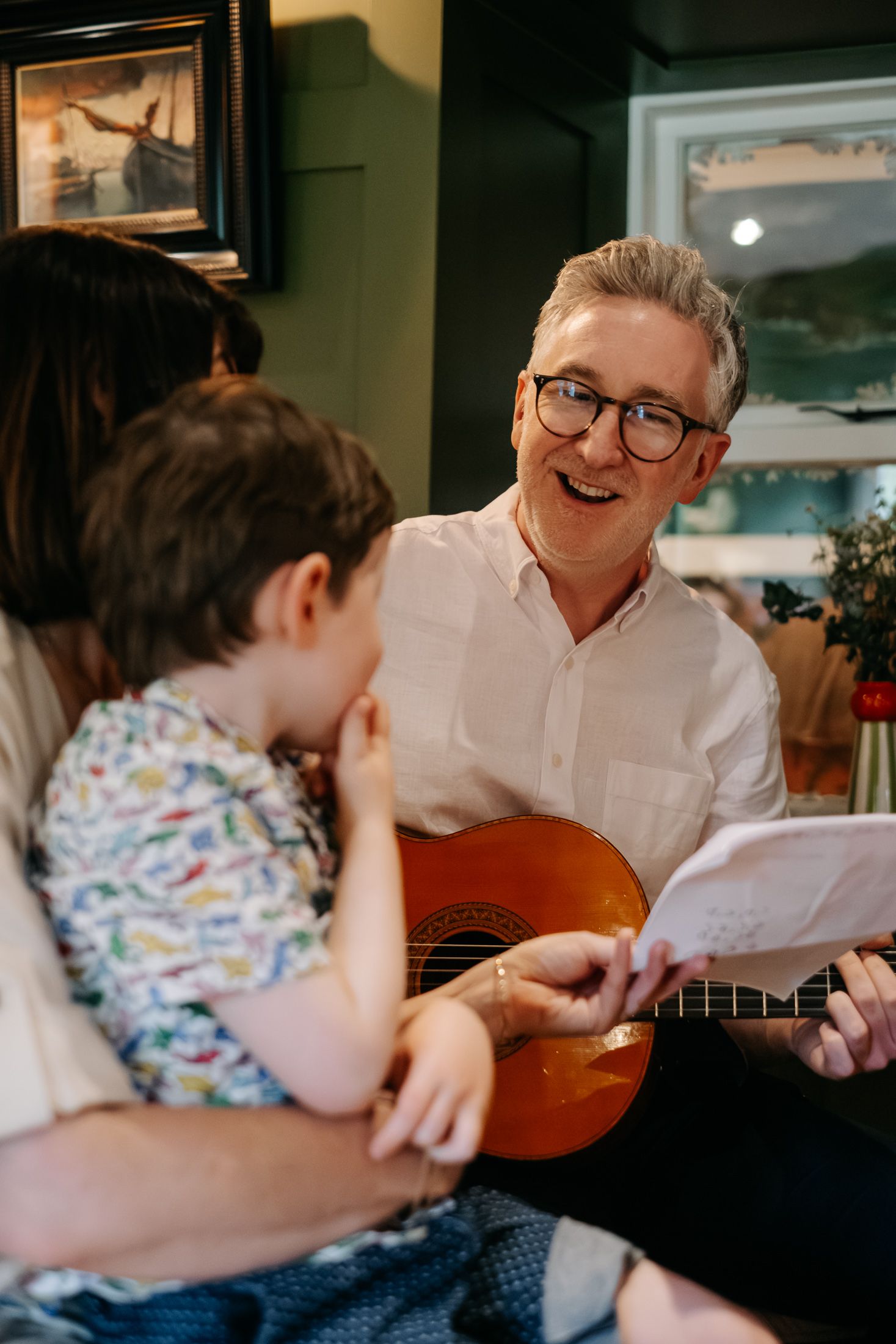 Smiling man with glasses plays acoustic guitar for a young child, holding sheet music indoors.