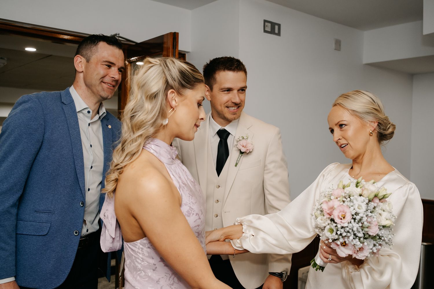Bride holding bouquet of pink flowers greeting guests in elegant wedding attire at indoor ceremony venue.