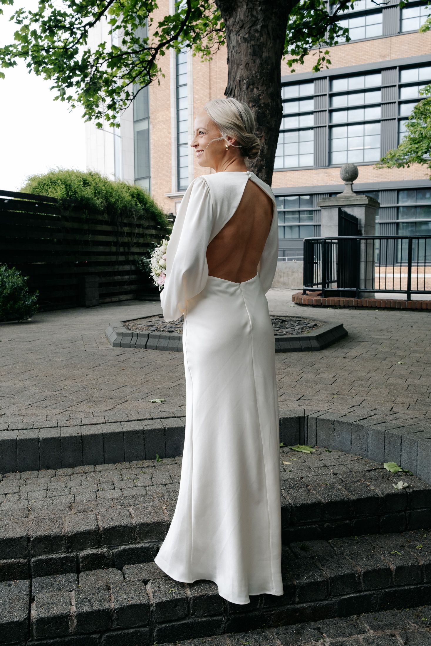 Woman in elegant white backless long-sleeve wedding gown standing outdoors near a tree on cobblestone steps.
