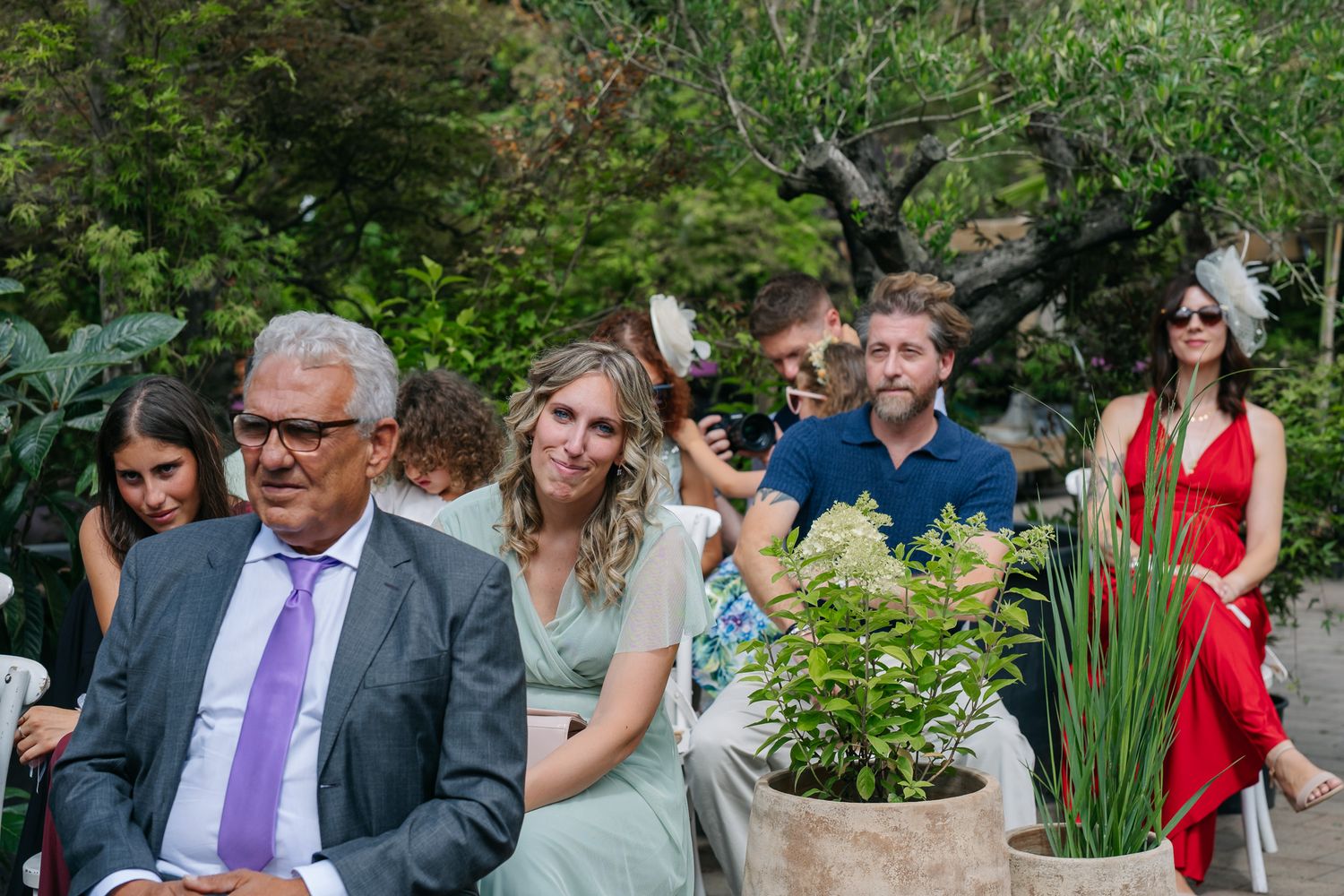 Guests seated outdoors at a garden wedding ceremony, with lush greenery and potted plants in the background.