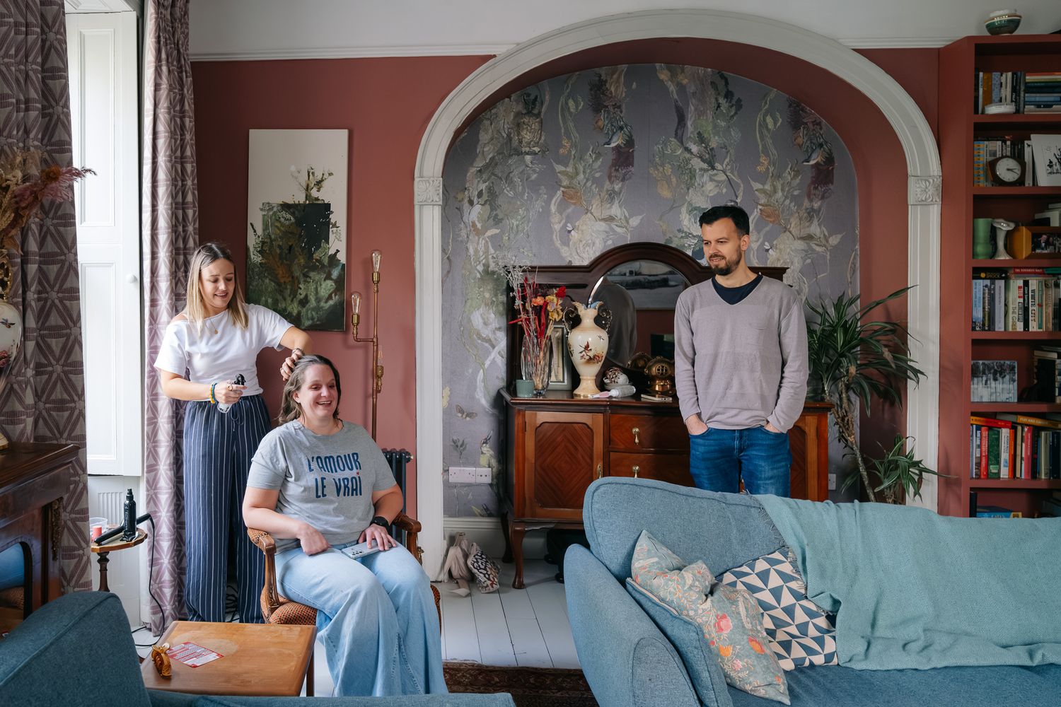 Three people pose in a stylish living room with blue sofa, pink walls, arched alcove, and decorative wallpaper mural.