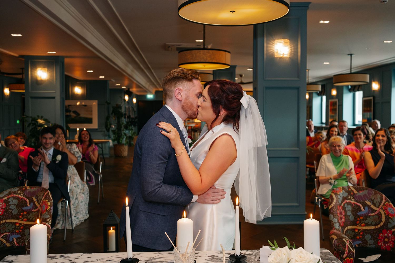 Wedding ceremony first kiss moment in a venue with teal columns and warm lighting.
