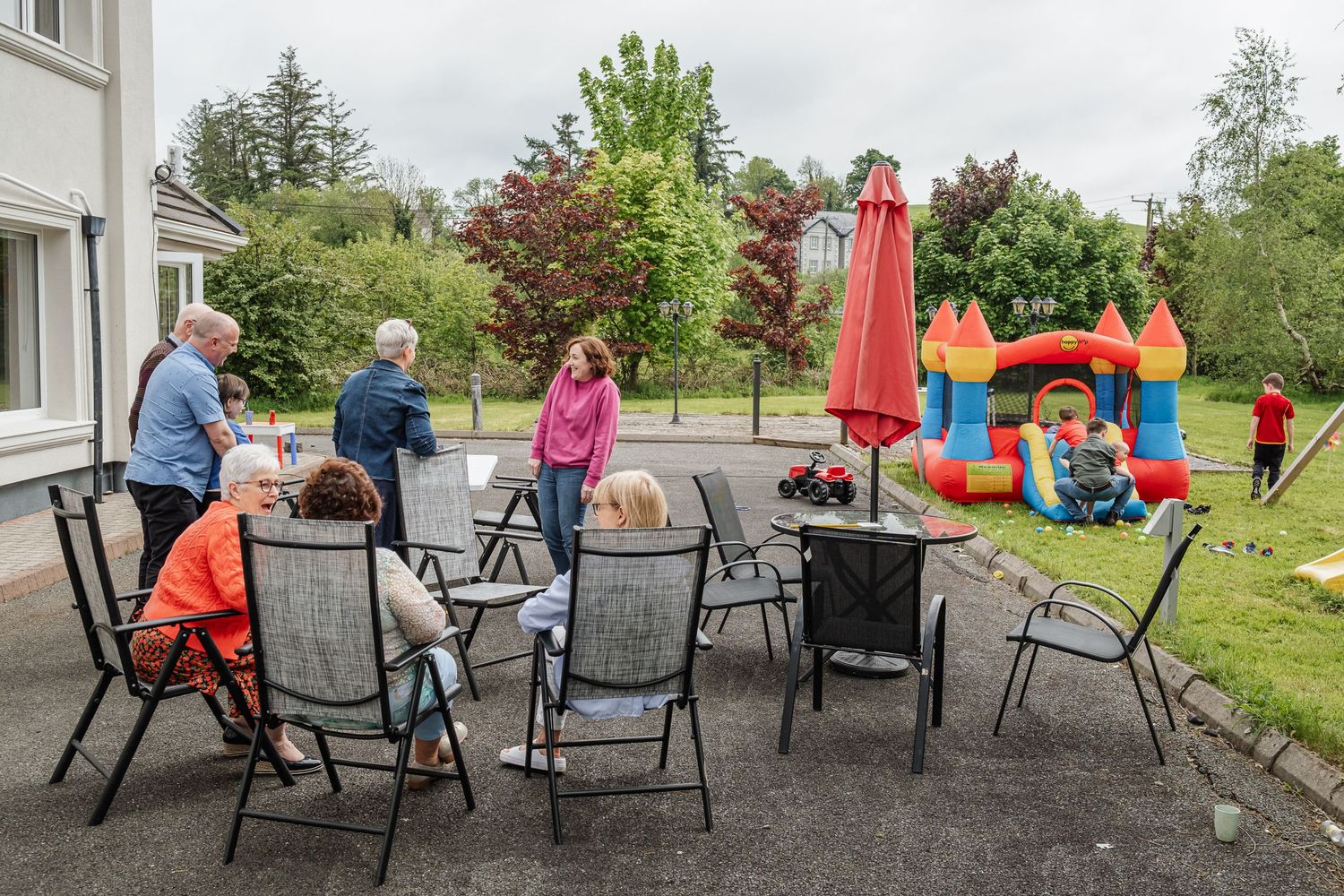 Adults sitting at patio furniture while children play on an inflatable bounce house in the background.