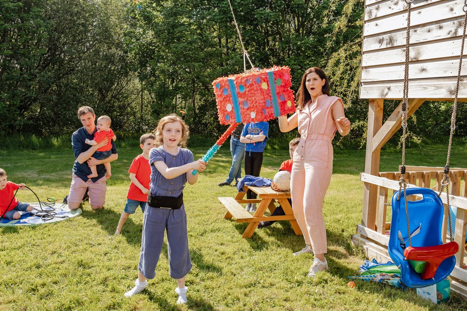 Children playing with a pinata in a sunny backyard with playground equipment.
