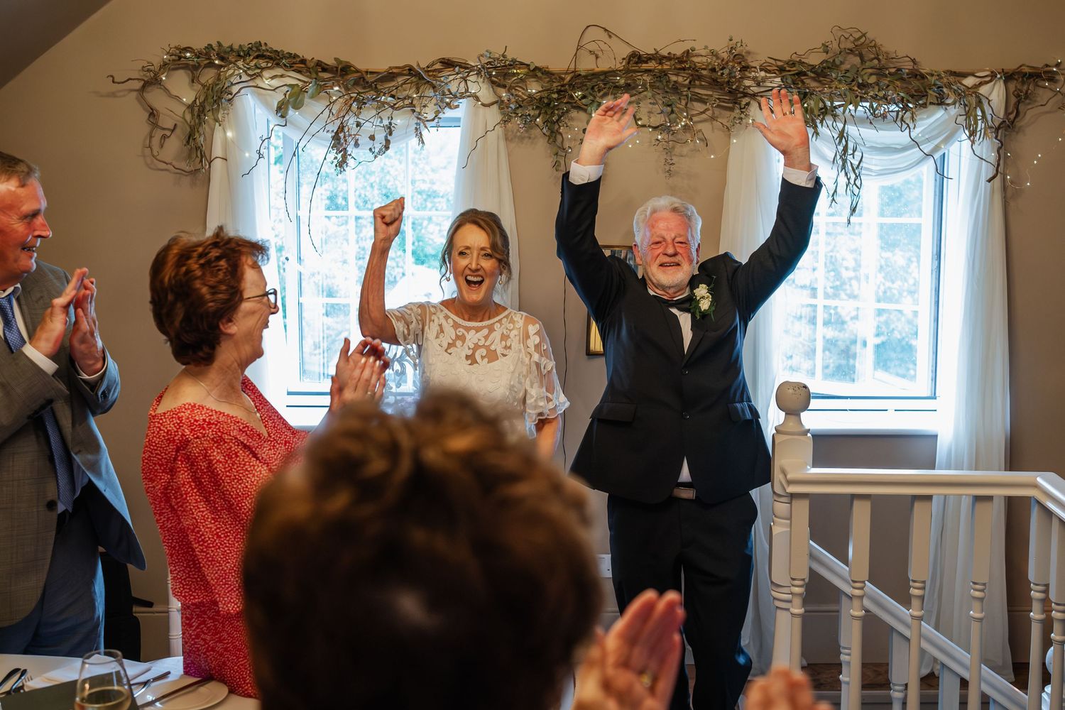 Wedding guests celebrate with raised hands under a window decorated with greenery garlands.