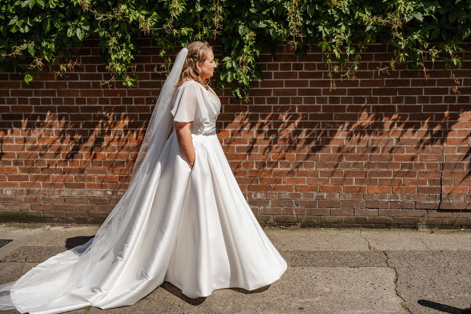 Bride in elegant white dress with flutter sleeves poses against brick wall with ivy.