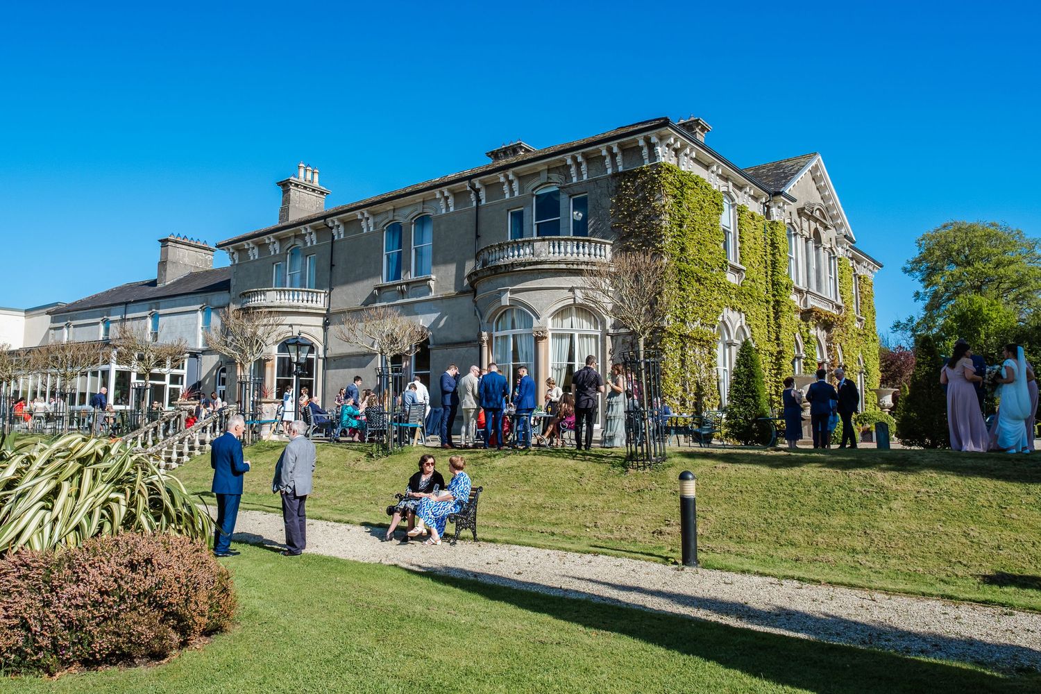 A grand stone mansion sits atop a grassy hill with people gathering outside on a sunny day.