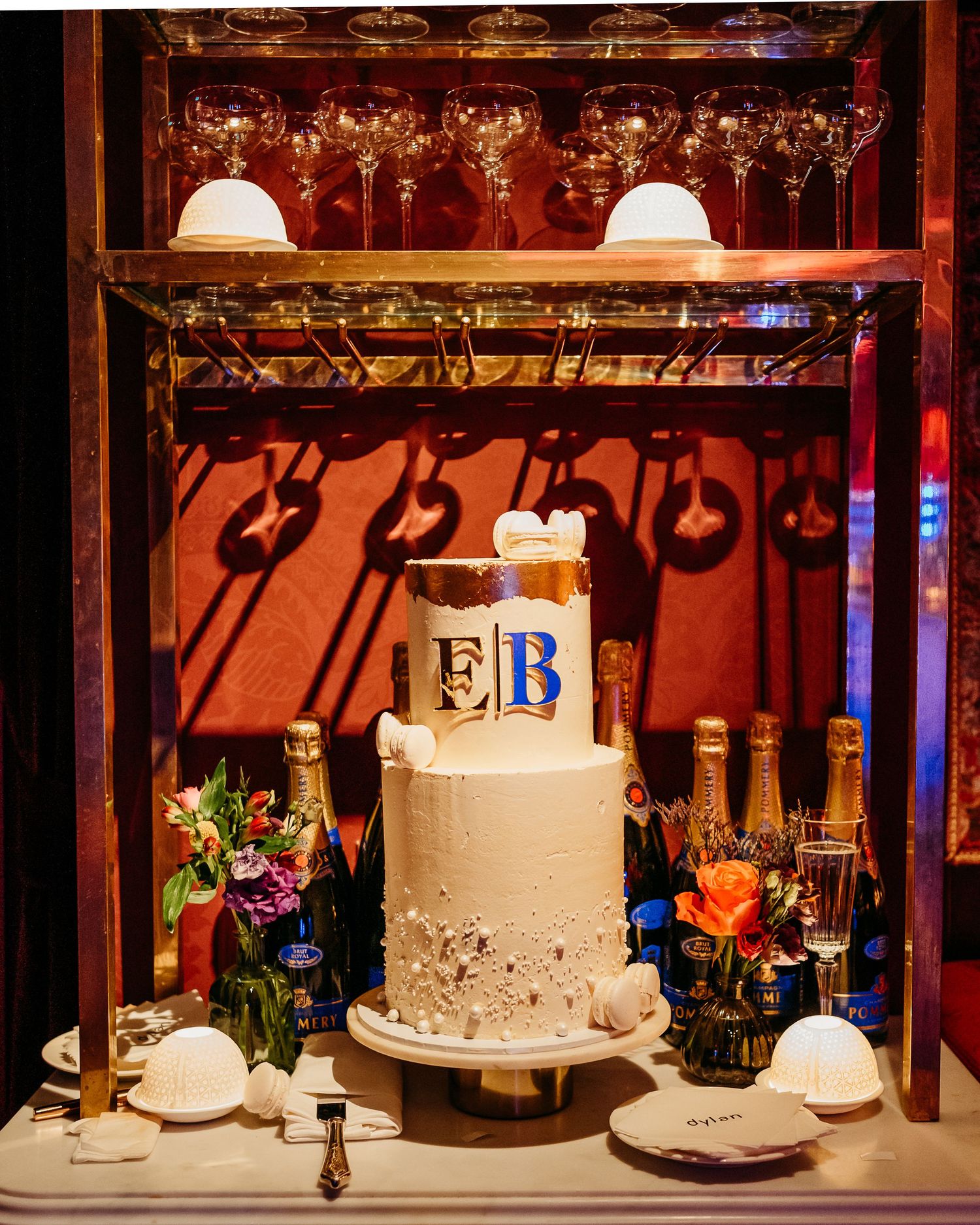An elegant white wedding cake with monogram decorations displayed on a bar shelf with ambient lighting.
