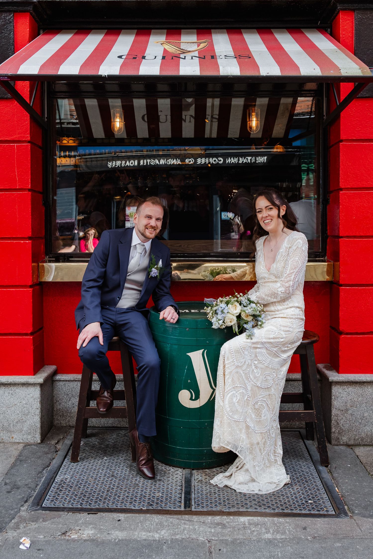 Newlyweds sitting on stools outside a red and white striped storefront in urban setting.