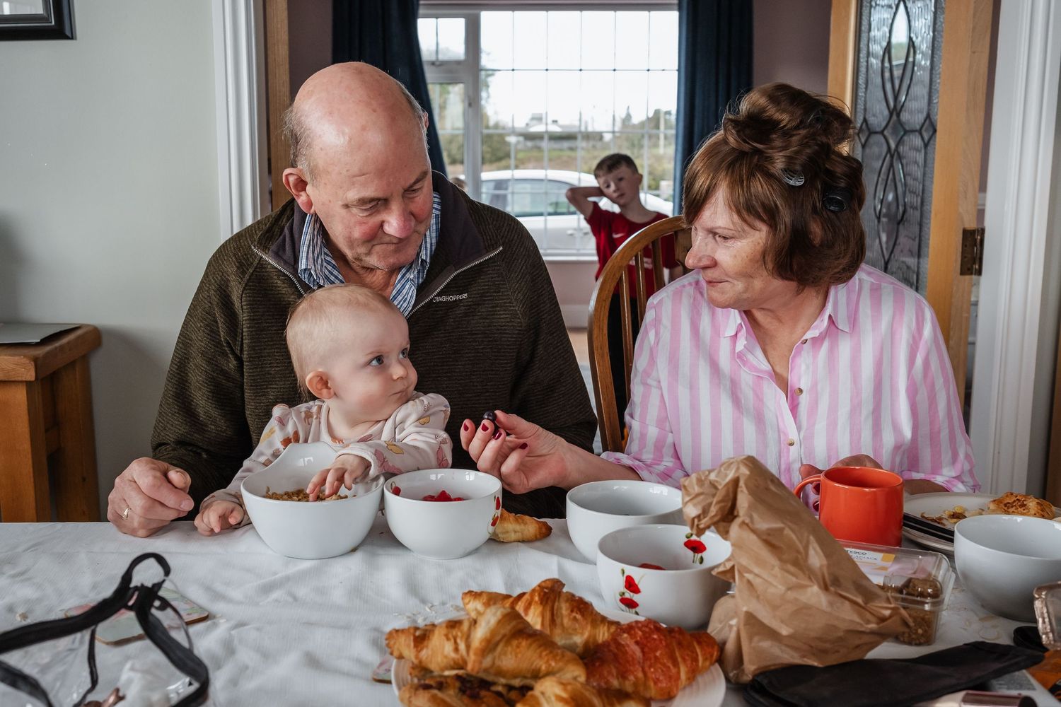Grandparents sharing a meal with a baby at a dining table, enjoying croissants and soup together indoors.