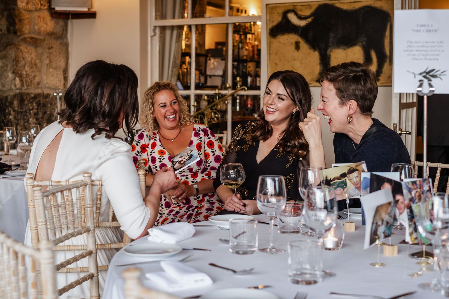 Four women laughing and enjoying wine together at an elegantly set dining table in a restaurant.