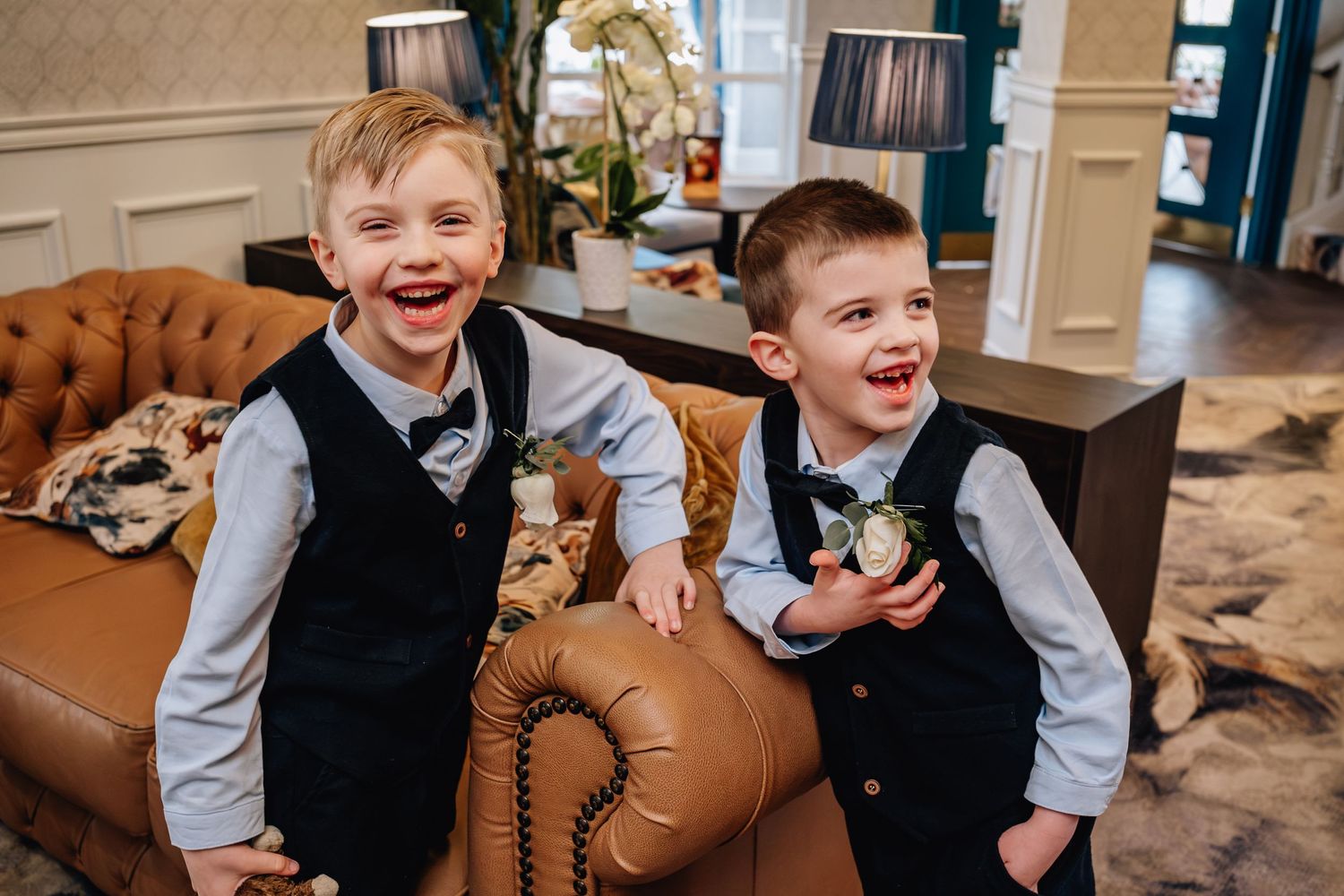 Two young pageboys in matching vests laughing on leather sofa.