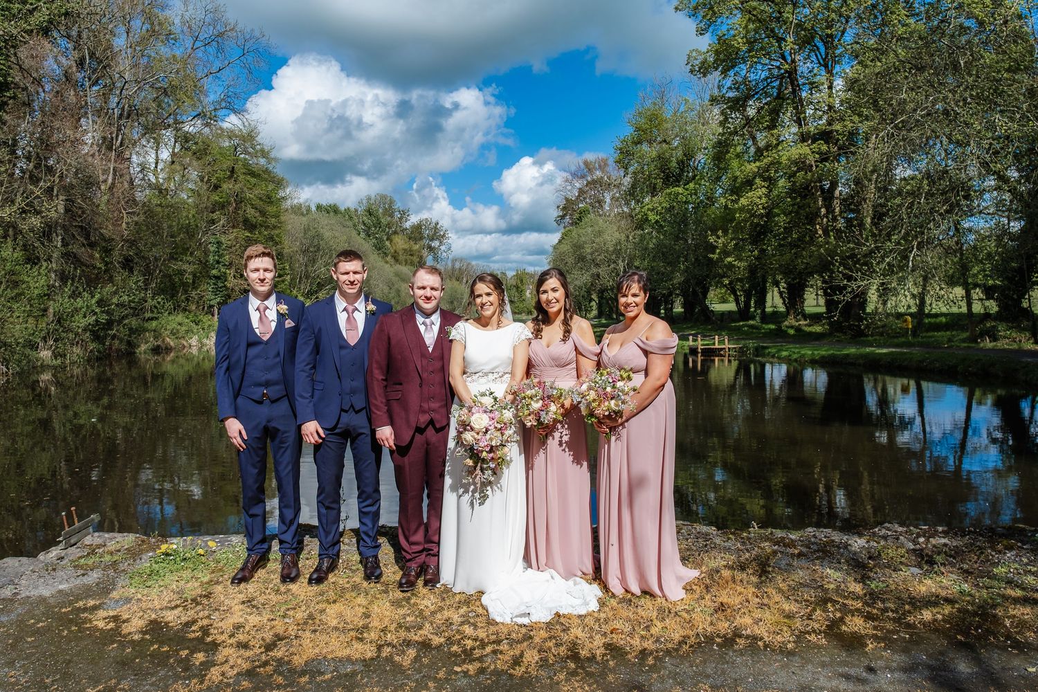Wedding party poses for group photos against scenic lake backdrop with blue sky and white clouds.