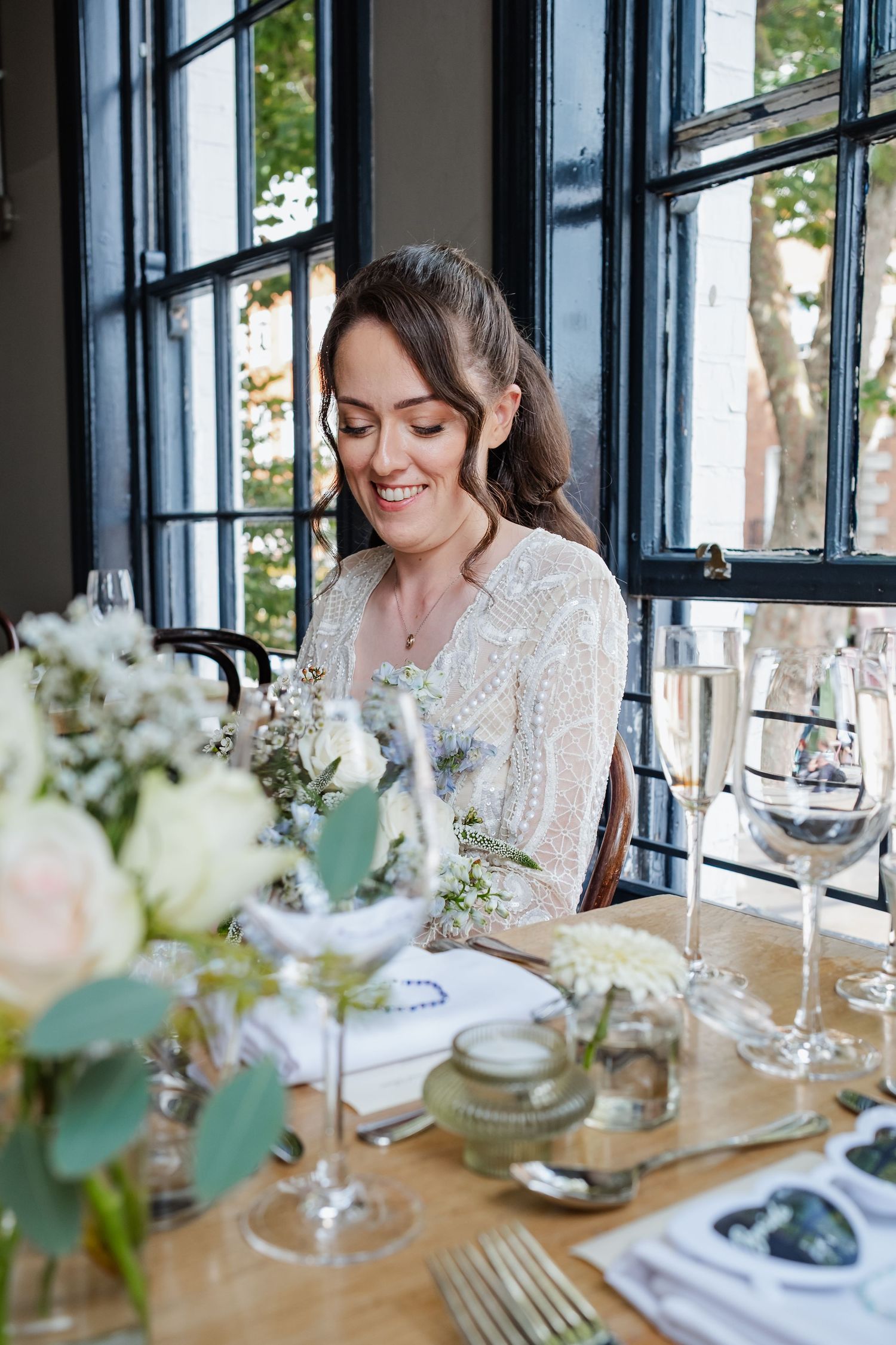 Wedding reception table setting with delicate floral centerpieces and wine glasses in a sunlit room.