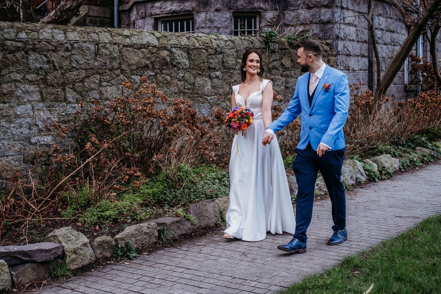 Bride and groom walk hand in hand on a stone path, smiling, with a rustic stone building backdrop.