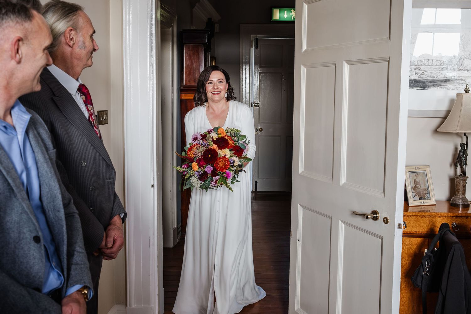 Bride in white gown holding colorful bouquet enters room as guests look on during wedding day preparations.