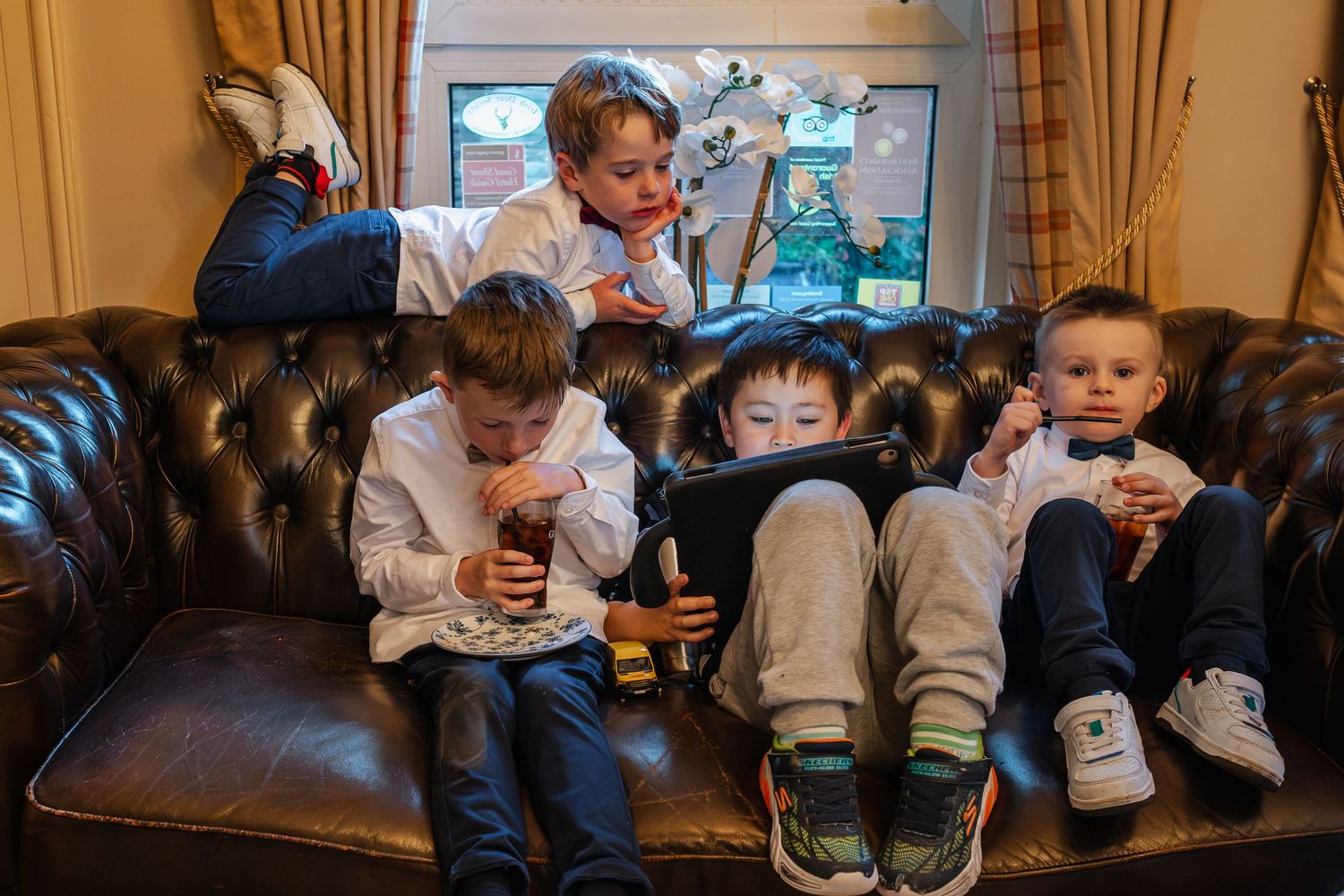 Four young boys relax on a leather sofa, watching a tablet and enjoying drinks at a family event.