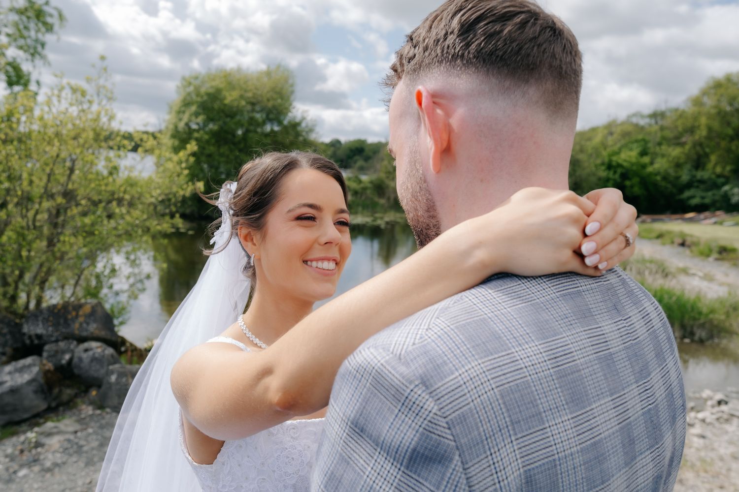 Smiling bride in white veil embraces groom in plaid suit outdoors near a lake on their wedding day.