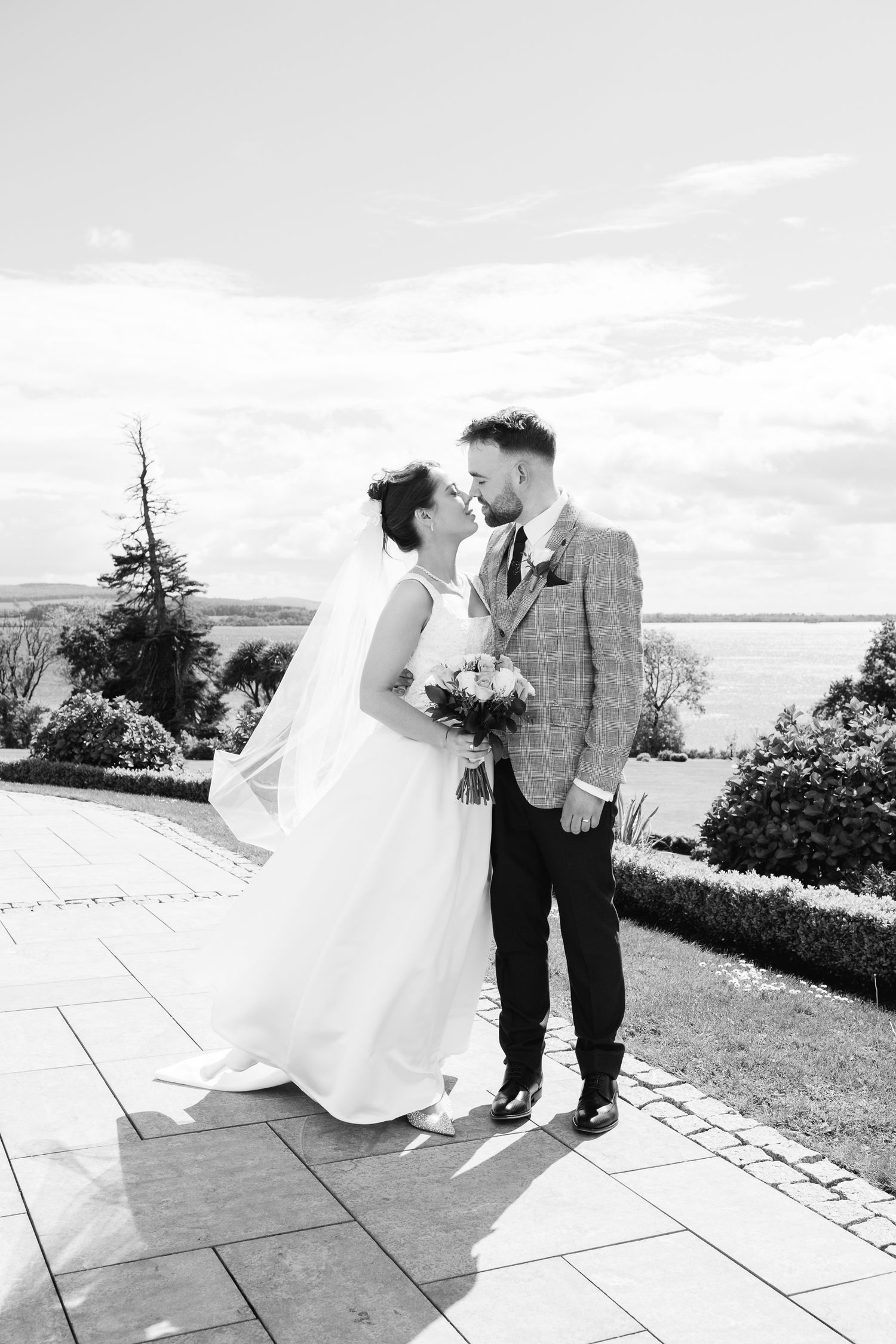 Black and white photo of a bride and groom sharing a romantic moment outdoors with a scenic waterfront backdrop.