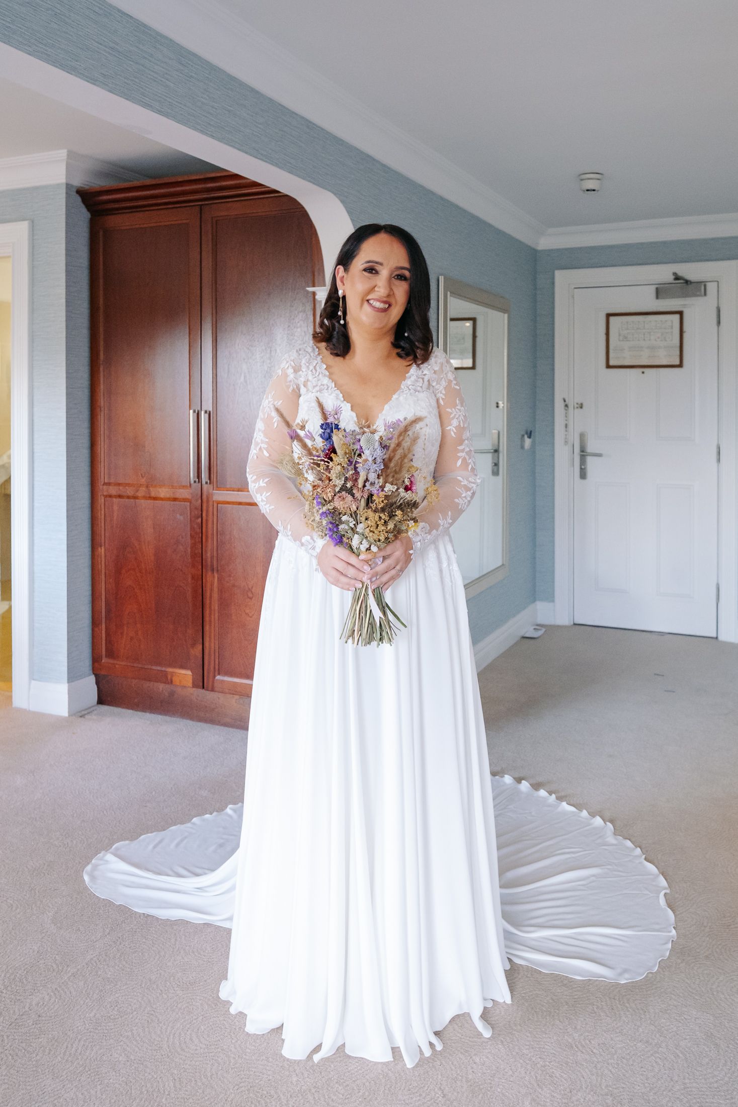 Smiling bride in lace long-sleeve wedding gown holding colorful wildflower bouquet in hotel room.