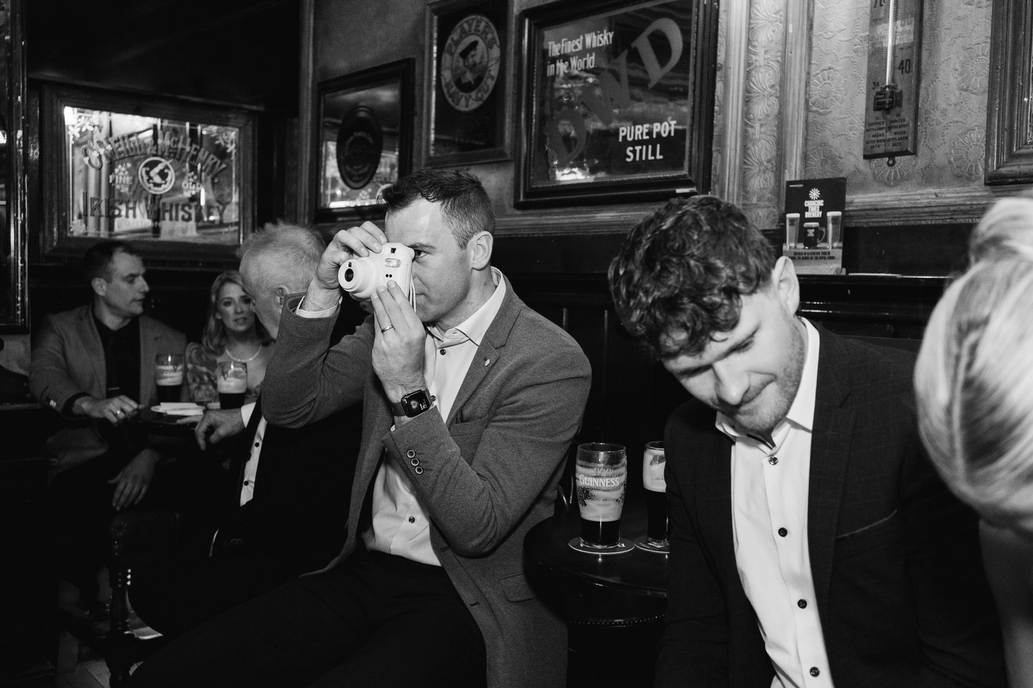 Black and white photo of wedding guests enjoying drinks inside a traditional Irish pub.