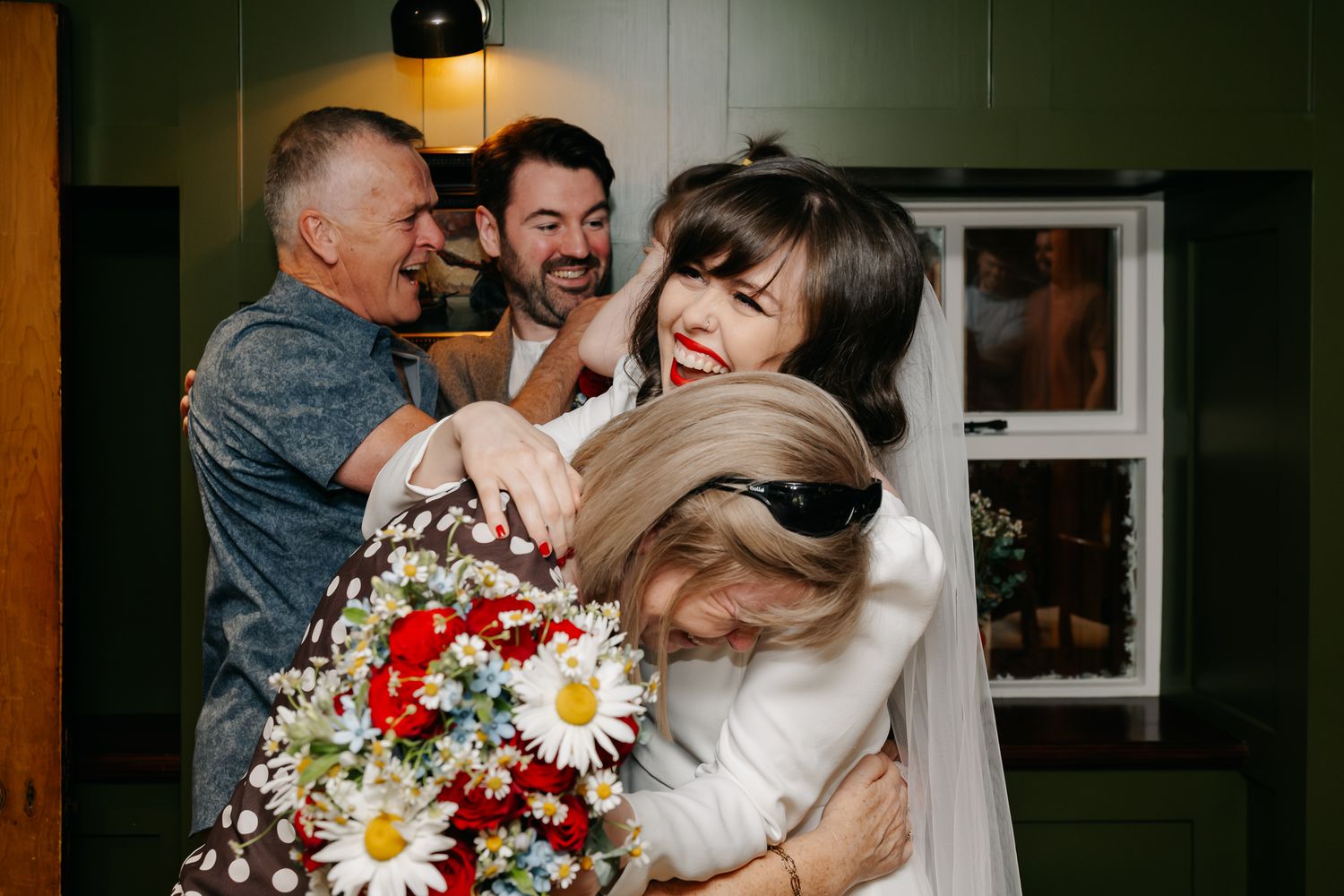 Bride in white dress hugging guests holding red and white flower bouquet at a joyful wedding celebration indoors.
