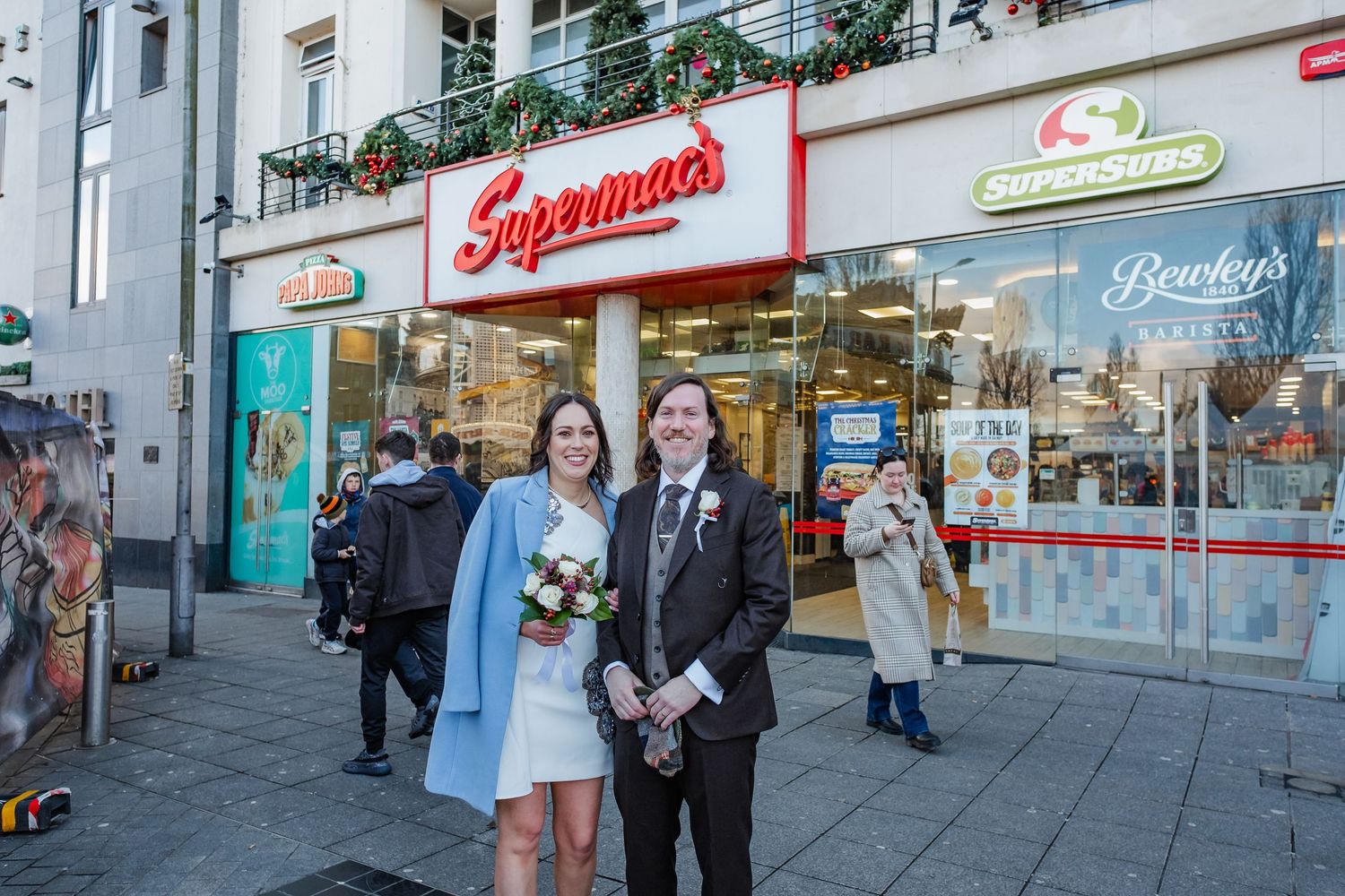 Newlywed couple poses in wedding attire outside Supermac's fast food restaurant on a city street.