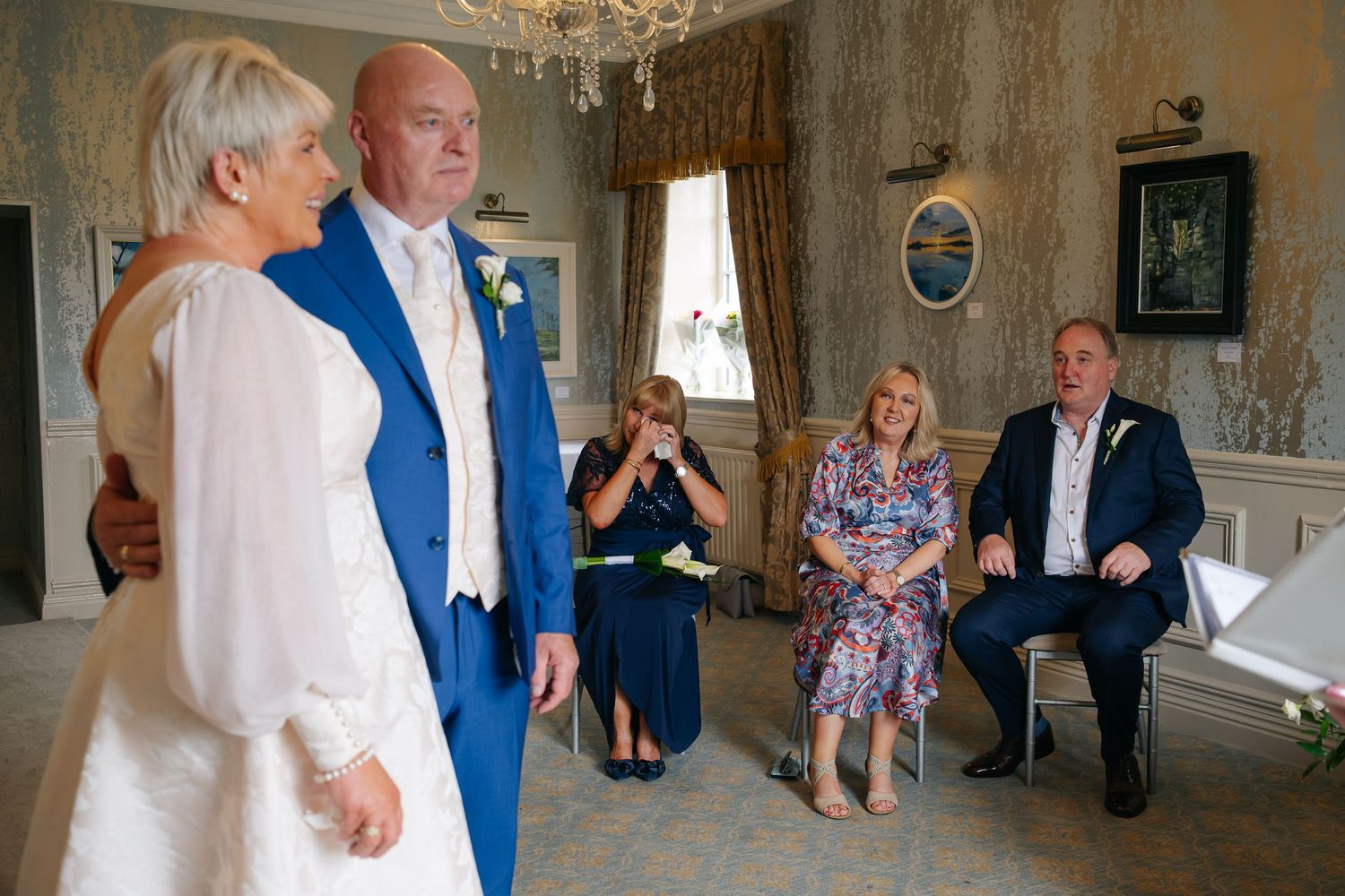 Mature bride and groom in blue suit exchange vows before seated wedding guests in elegant room.