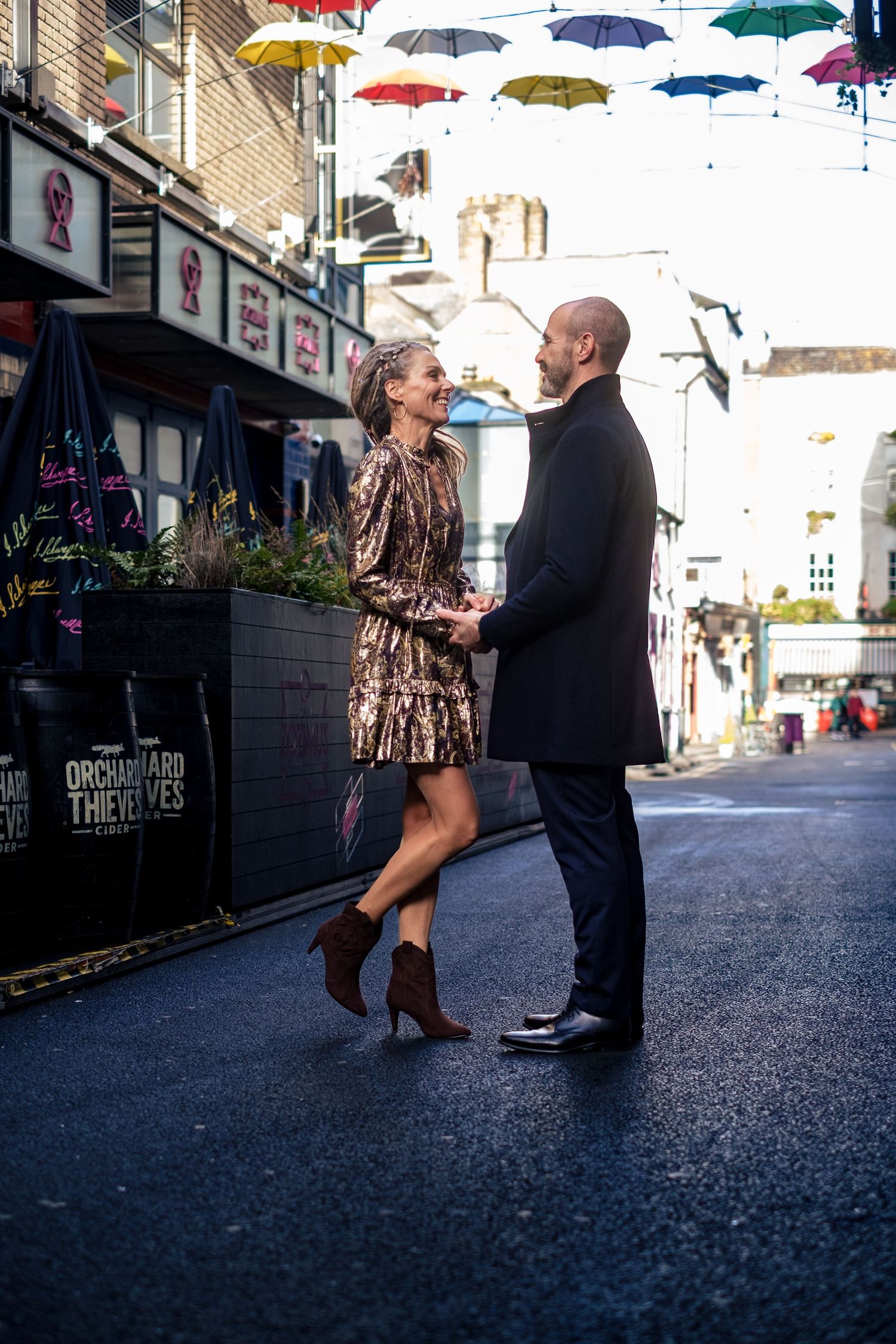Couple holding hands on a colorful umbrella-lined street, woman in gold dress and brown boots, man in dark suit.