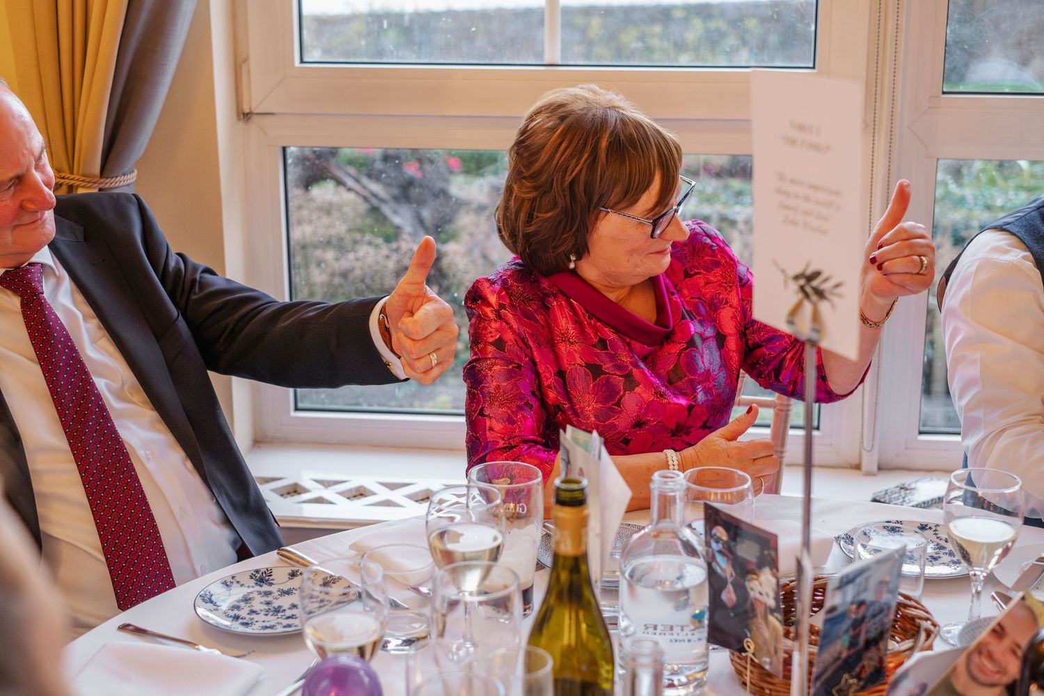Wedding guests in formal attire gesturing animatedly during a reception dinner with wine on the table.