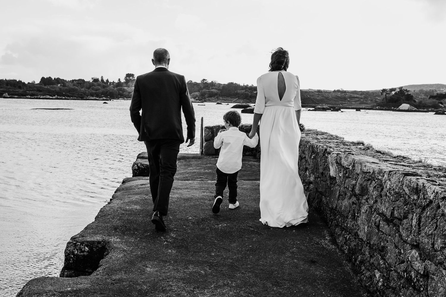 Black and white photo of a couple in wedding attire walking with a young child on a stone pier by the sea.