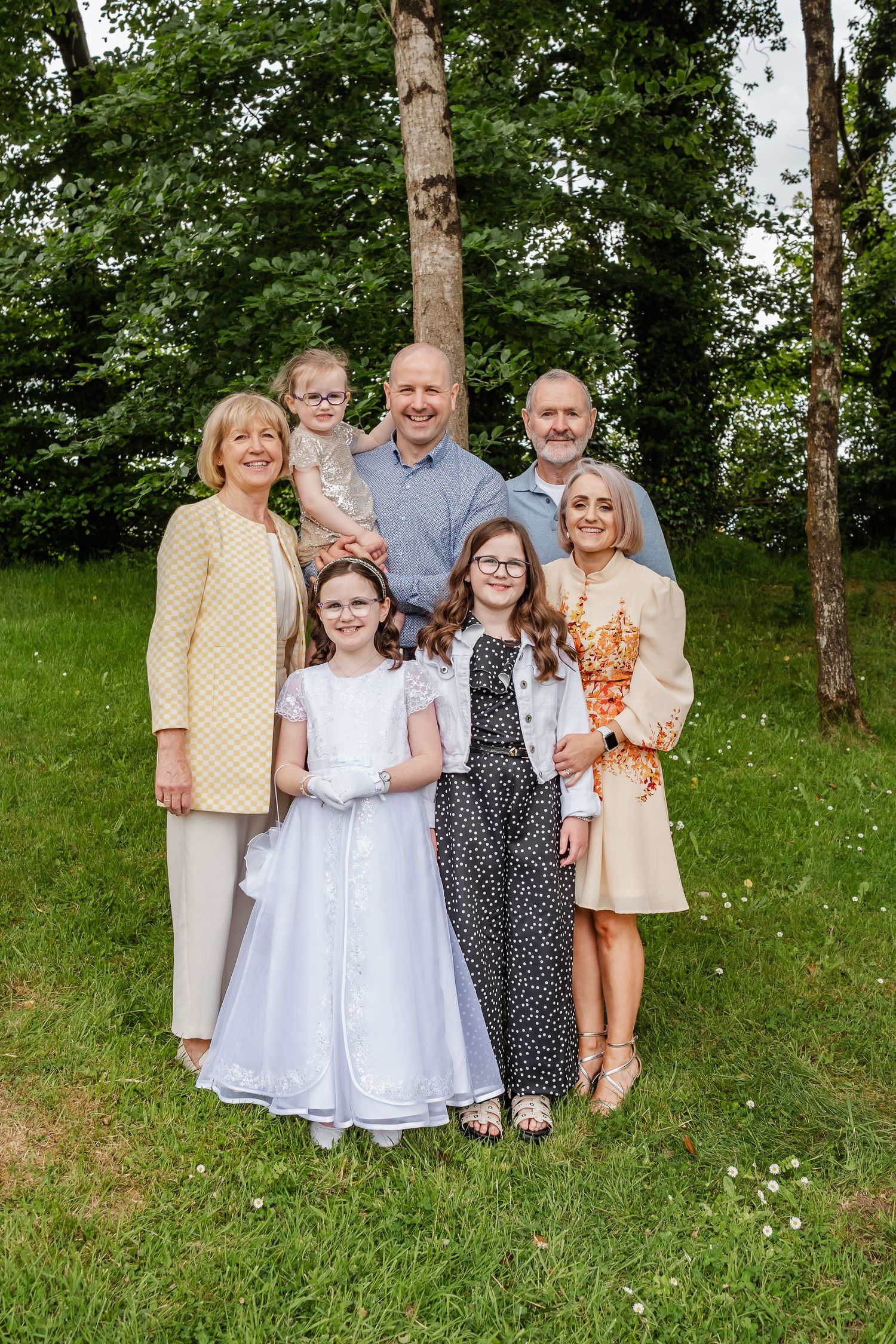Family portrait of seven people posing outdoors on green grass with trees in background, including girls in white communion dress.