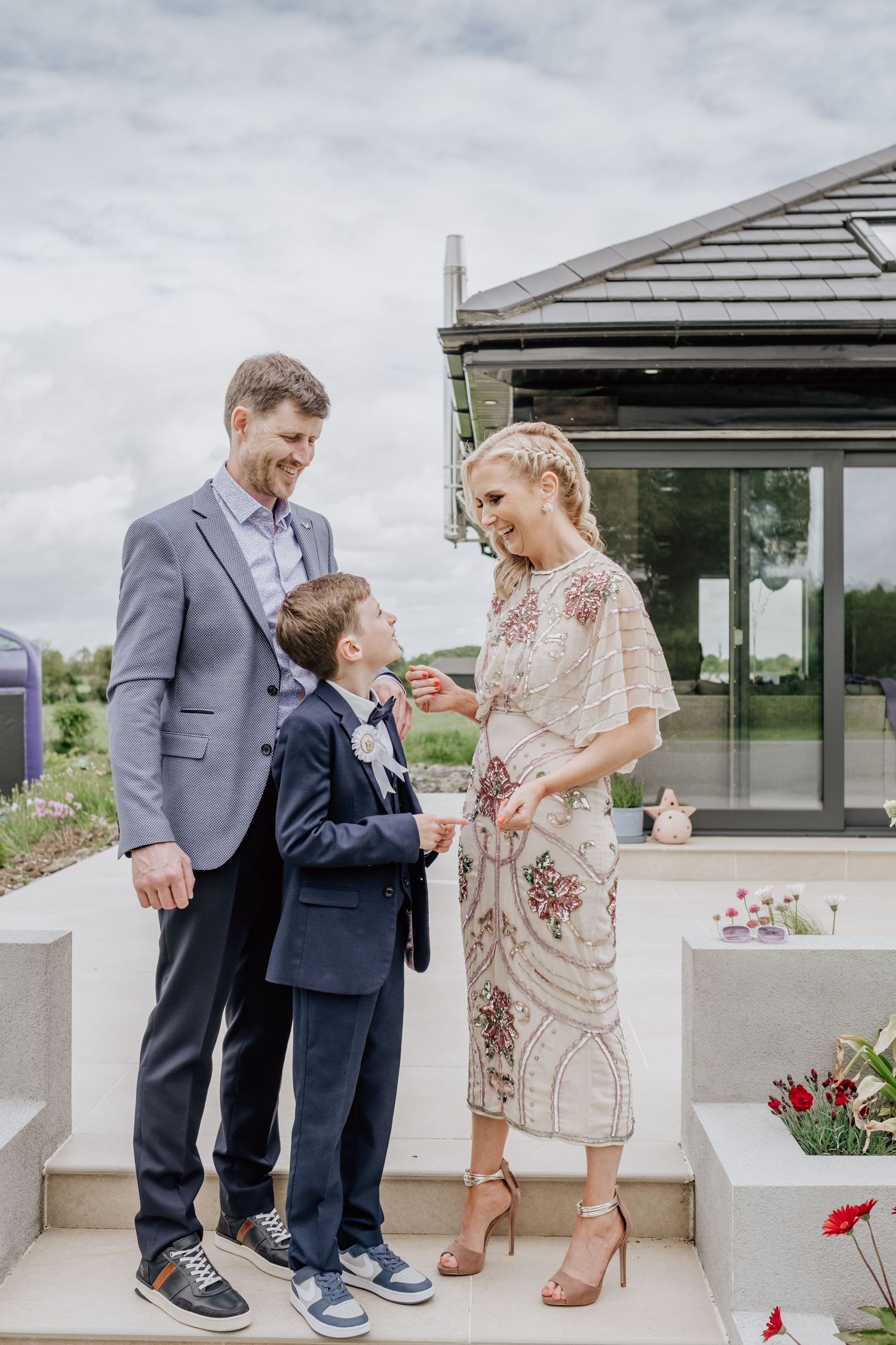 A family of three in formal attire poses outdoors at a wedding venue, with a boy in a navy suit between his smiling parents.