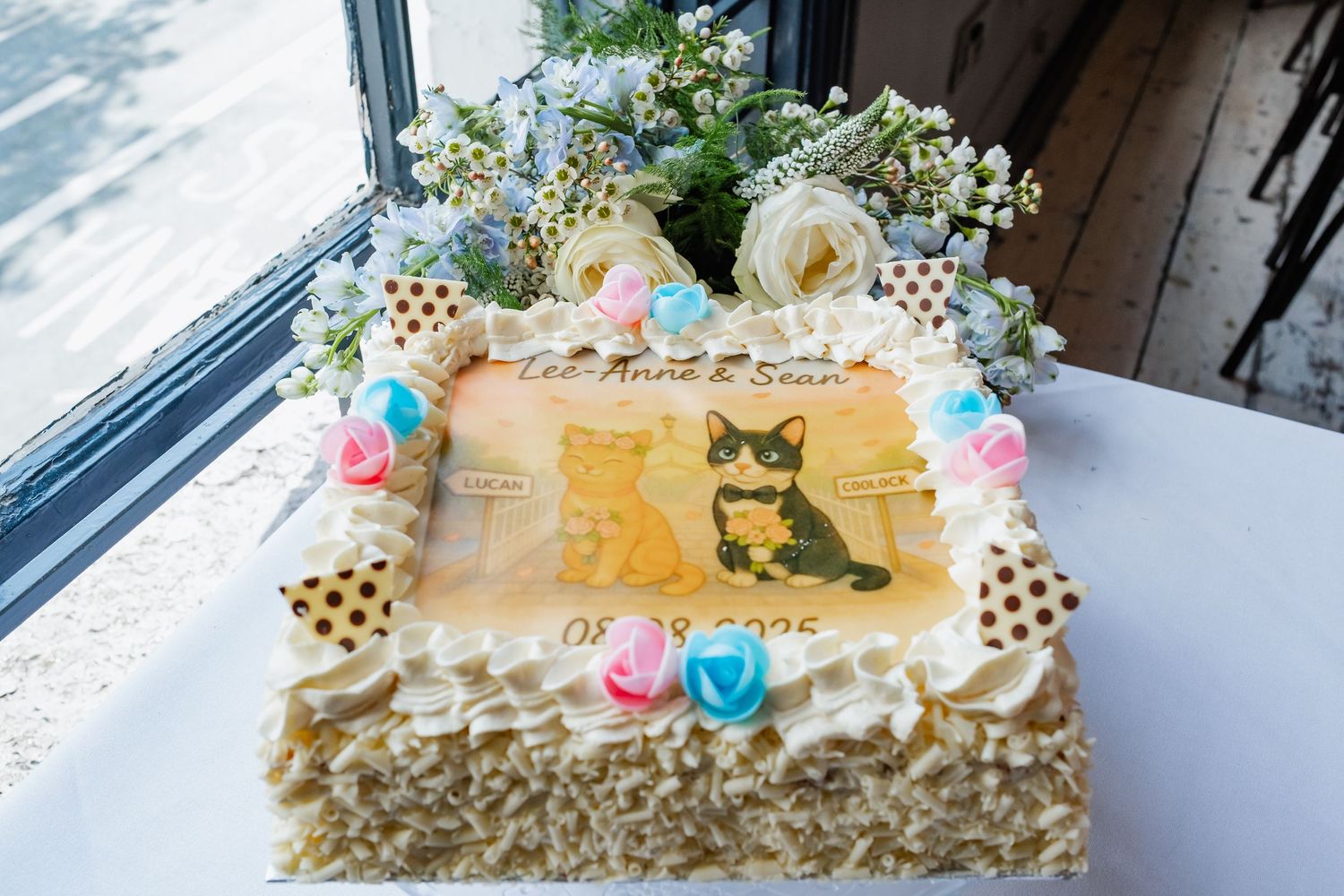 A decorated white cake with colorful frosting flowers and a cat figurine on top sits on a glass serving plate.