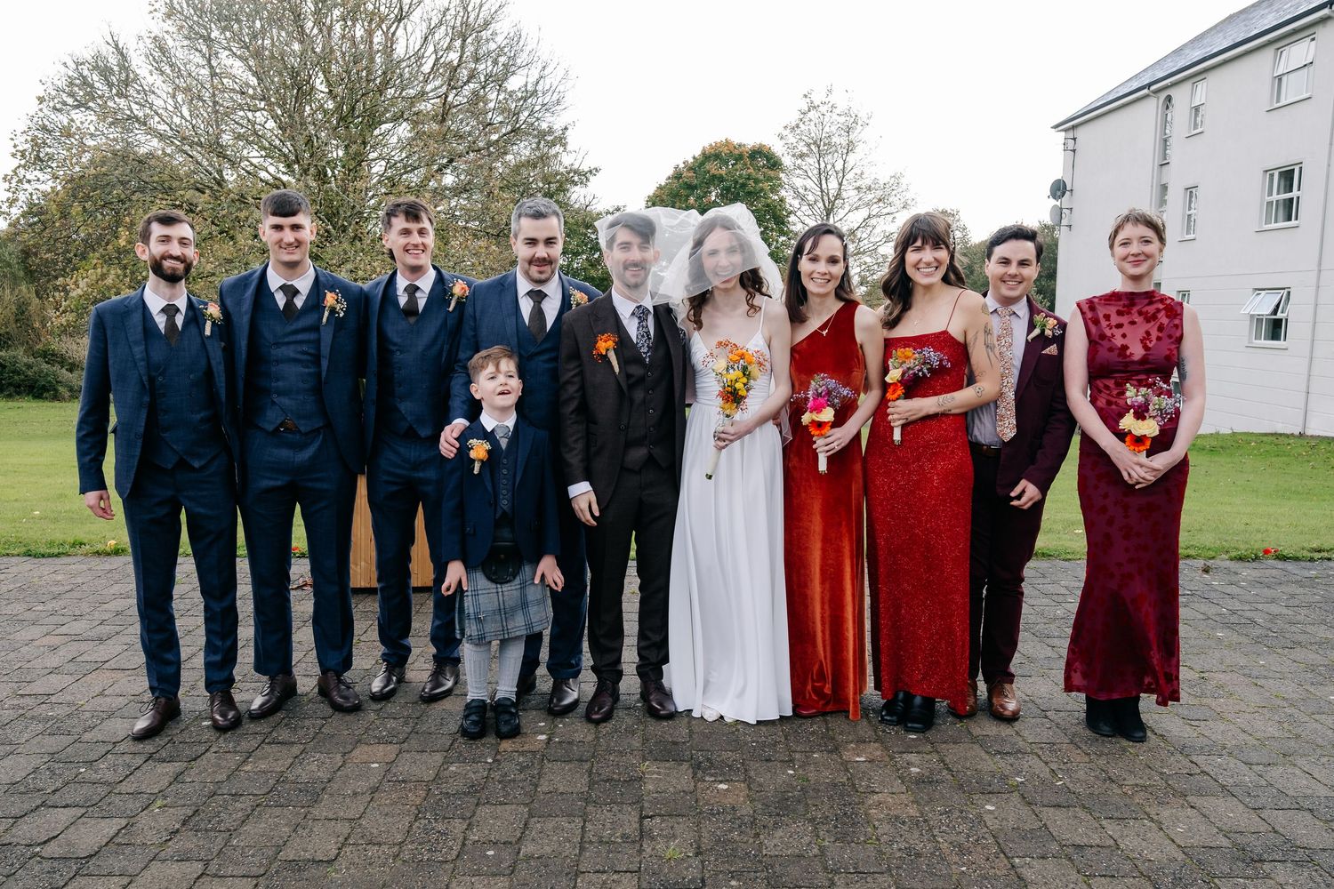 Large wedding party group photo with bridesmaids in red dresses and groomsmen in navy suits.
