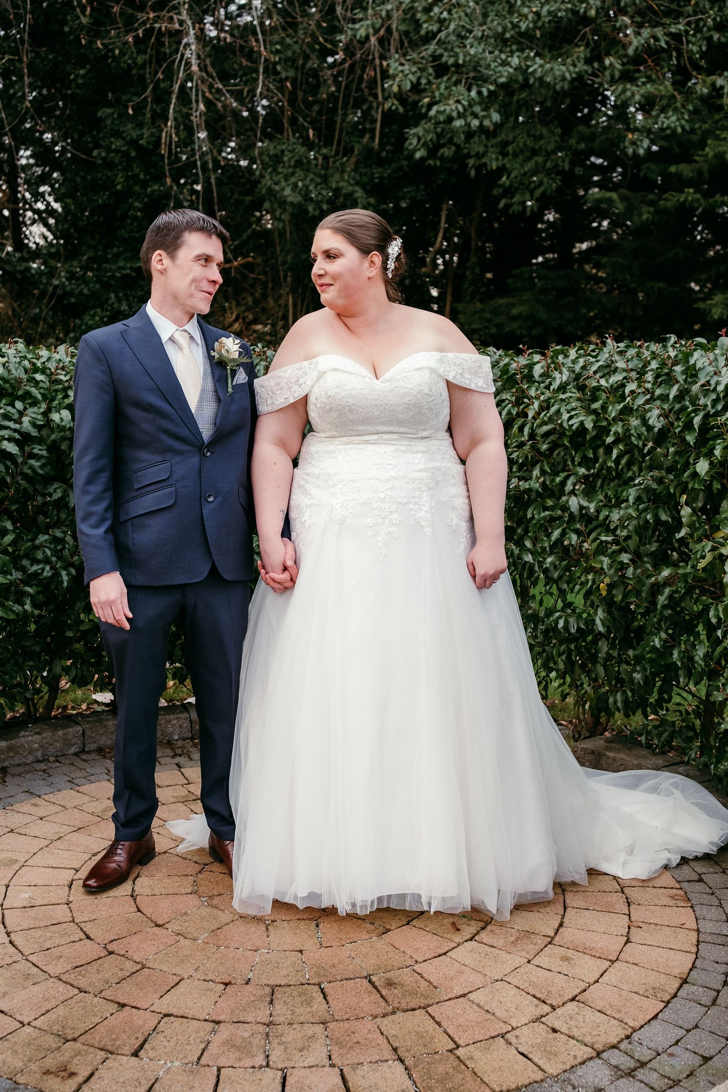 Wedding couple standing on a circular brick patio surrounded by lush greenery.
