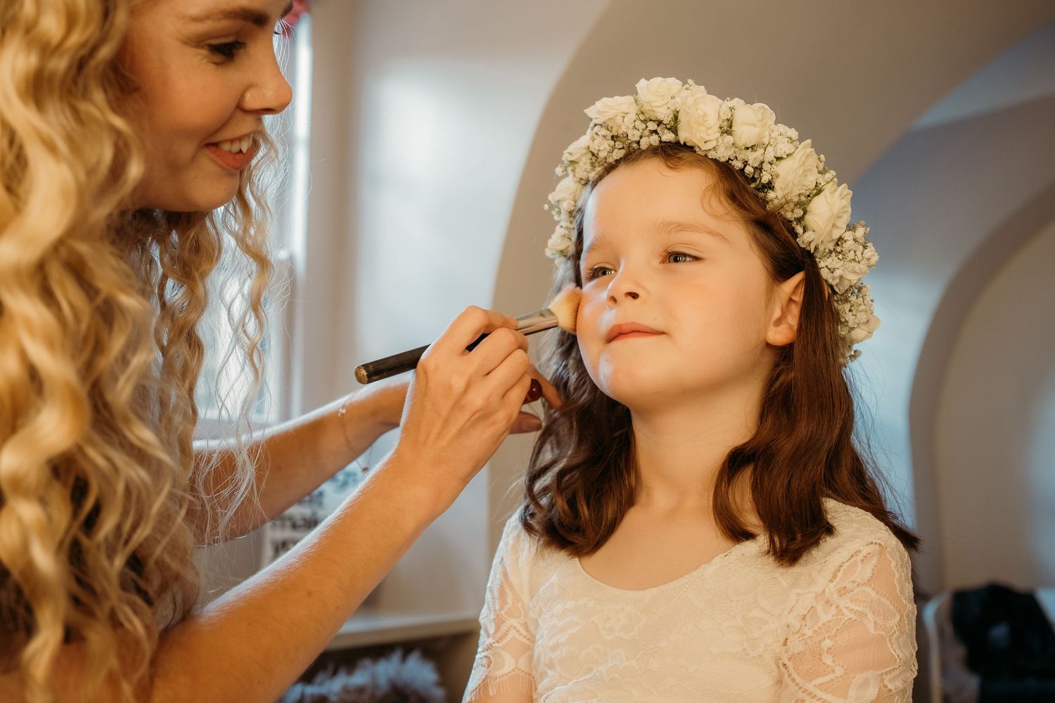 A stylist applies makeup to a young attendant wearing a white flower crown.